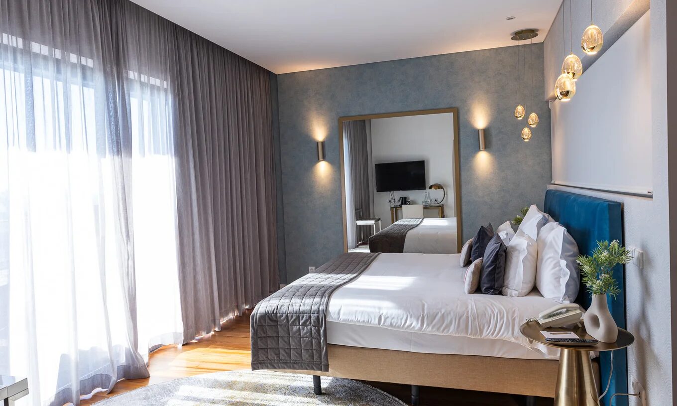 Quarto moderno com cama de casal, cortinas amplas, iluminação suspensa e espelho diante da televisão