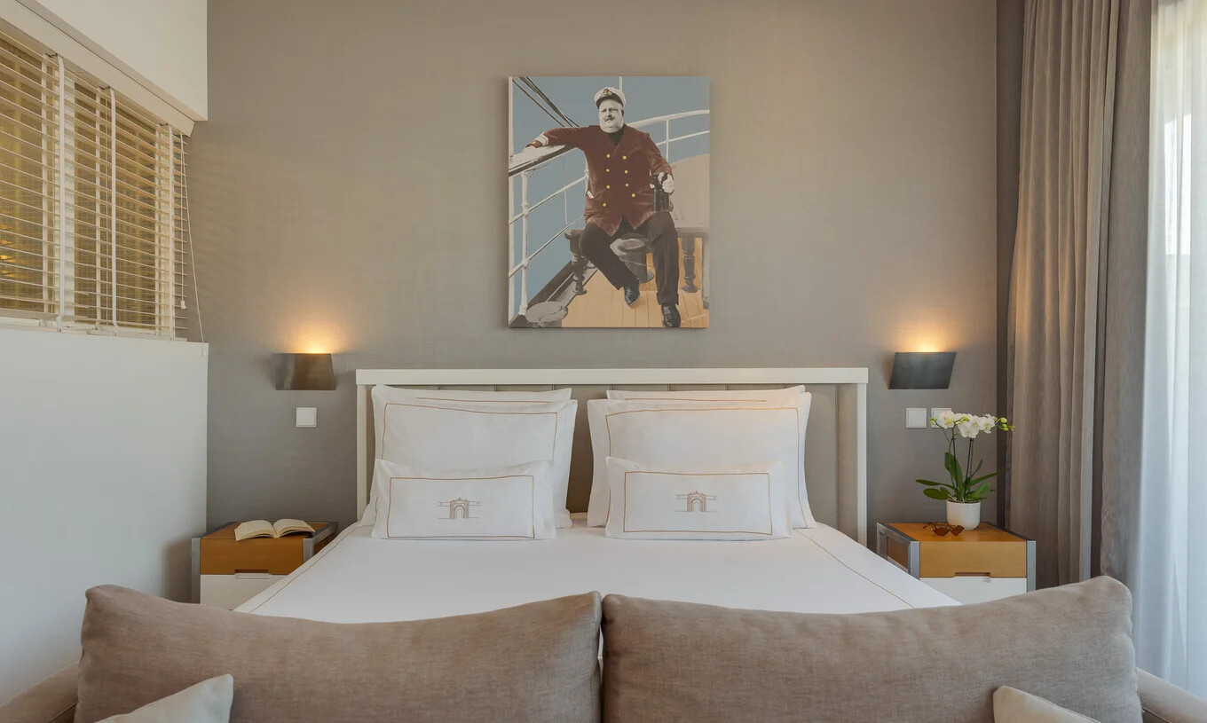 Quarto com cama de casal, iluminação suave, quadro sobre a cabeceira, cortinas e mesa lateral junto à janela