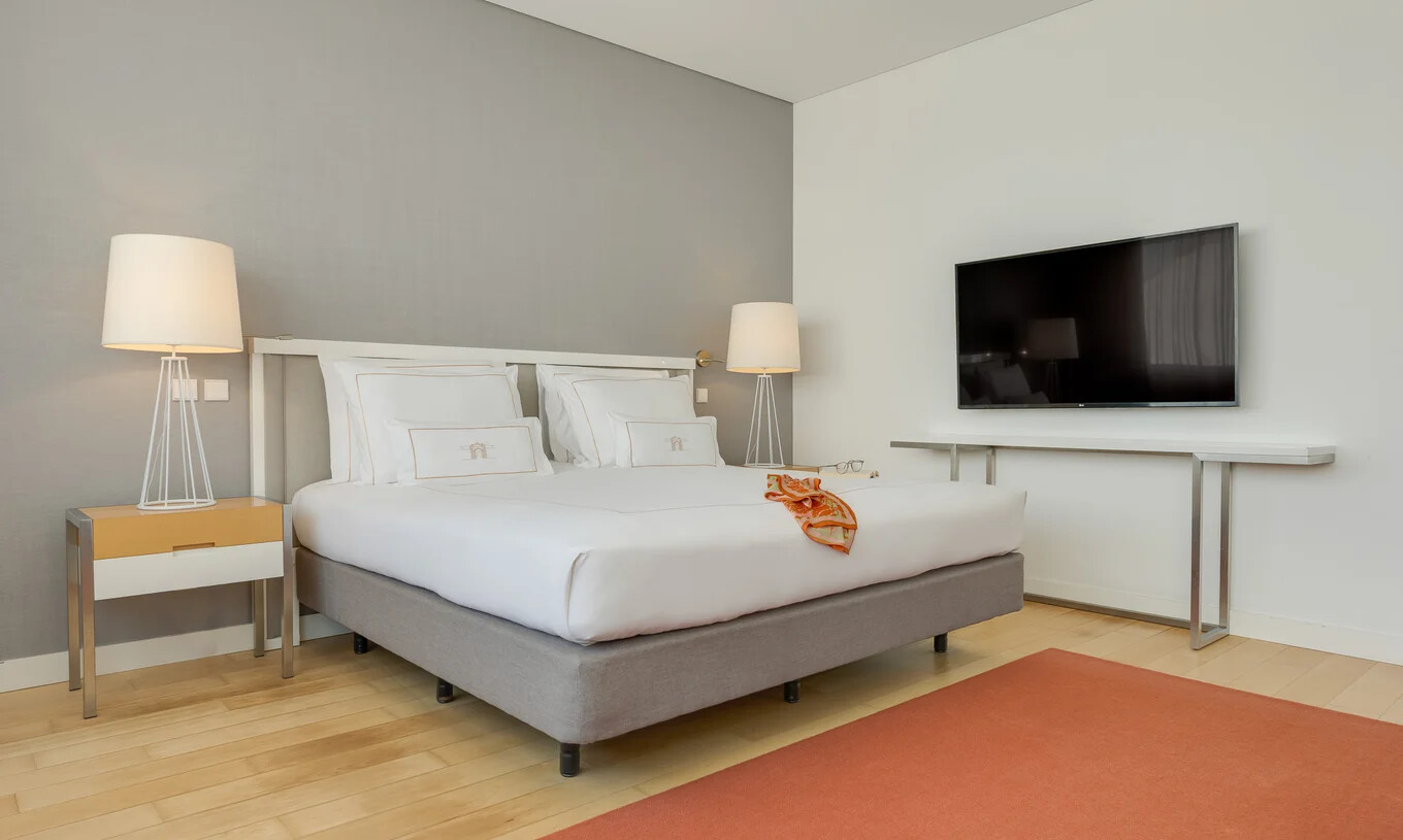 Quarto com cama de casal, televisão de parede, candeeiros altos e decoração minimalista em tons claros