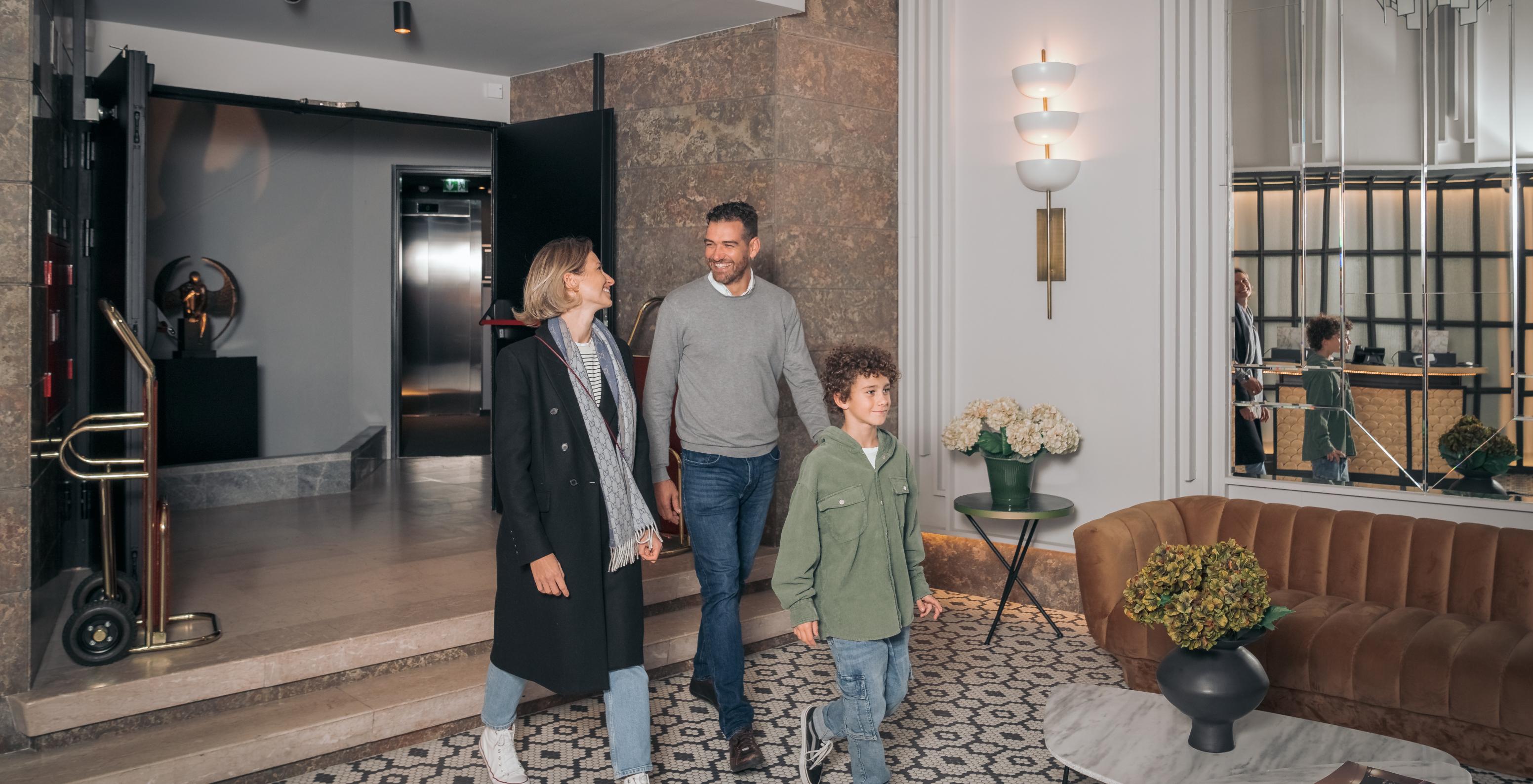 Lounge da receção com um casal e uma criança a andar, espelhos, um sofá e mesas de apoio com vasos de flores