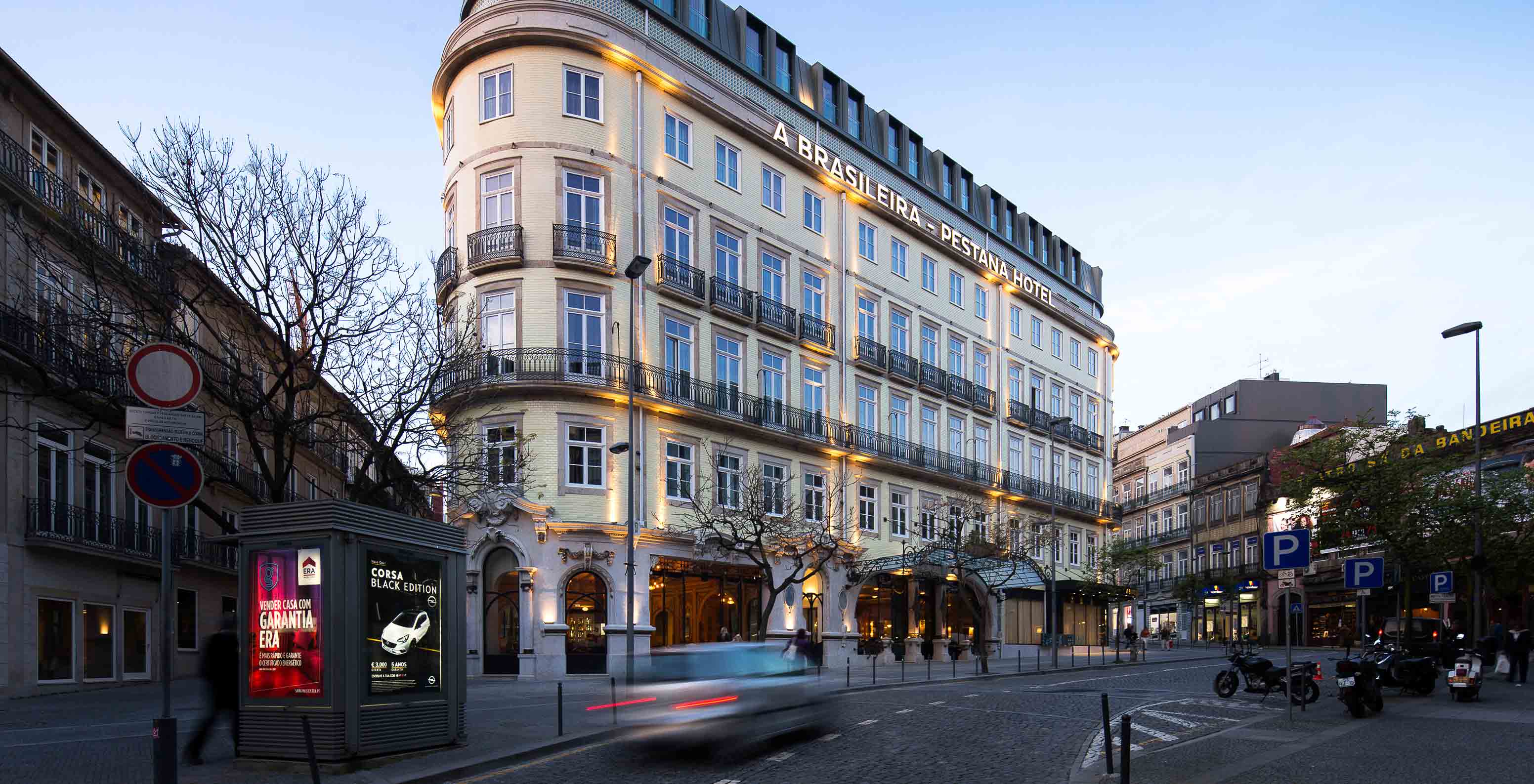 Fachada iluminada do Pestana Porto A Brasileira vista da rua com edifícios e árvores ao redor