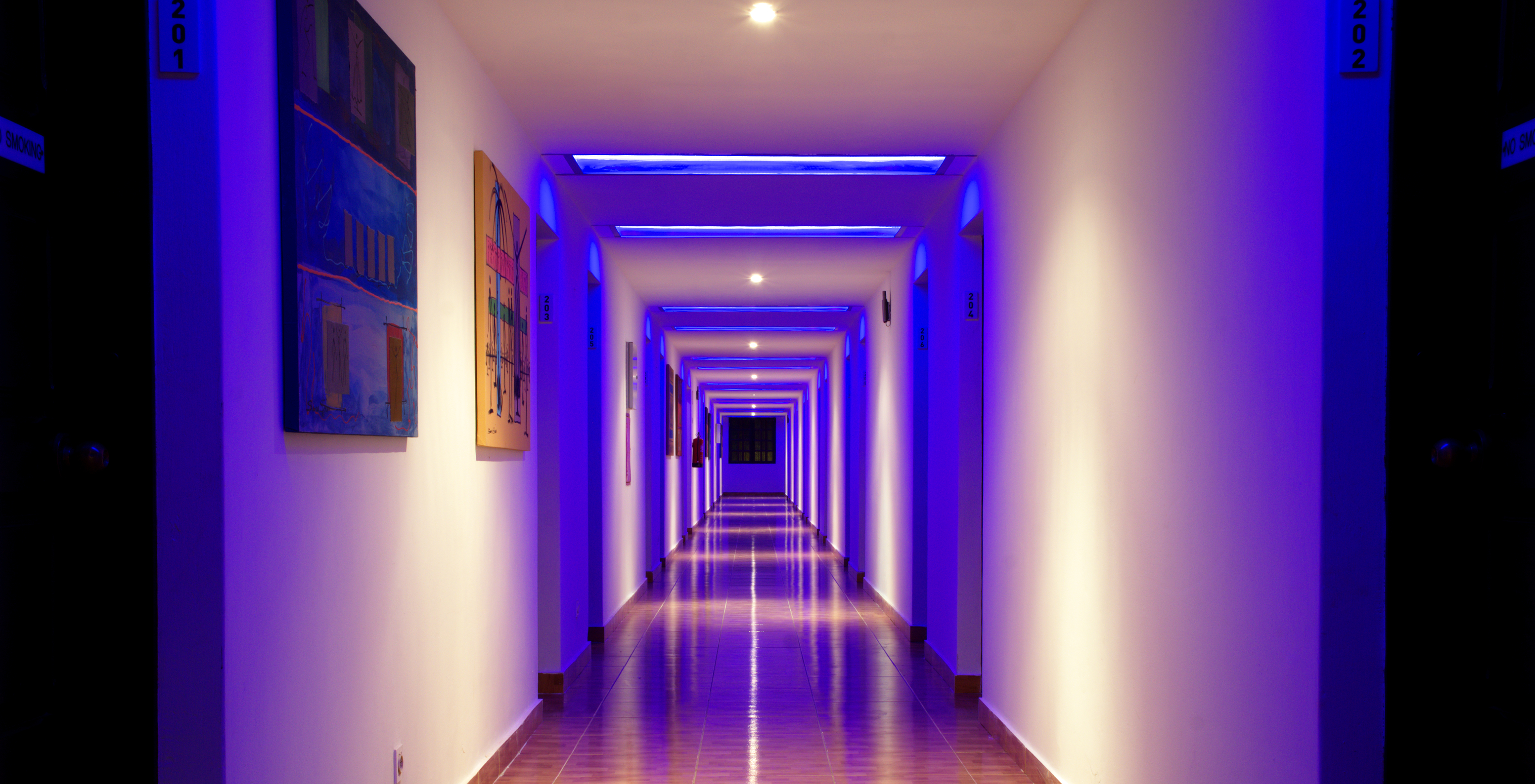 Corredor de quartos do Hotel com Piscina, em Frente à Praia, com luzes azuis néon e dois quadros coloridos