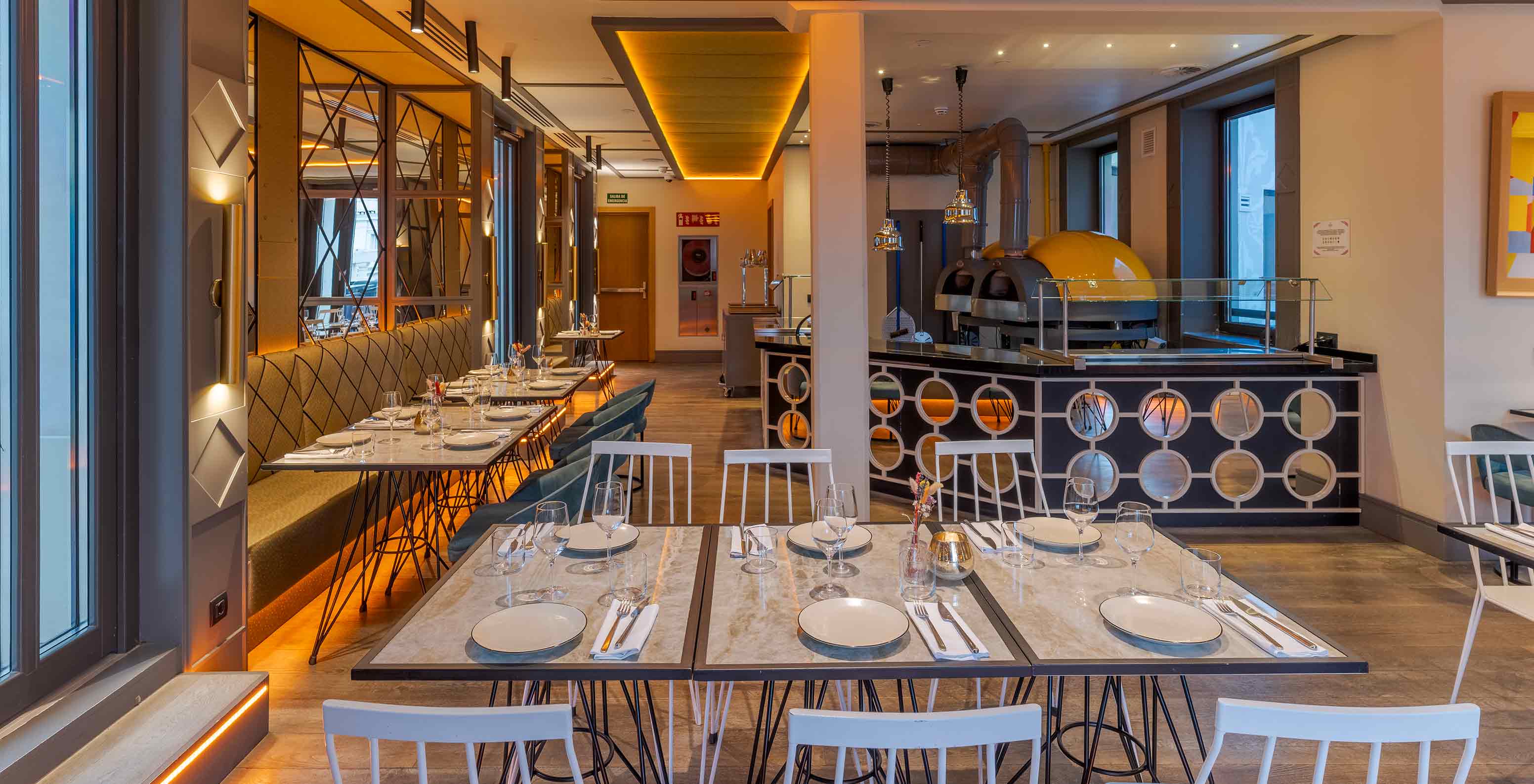 Interior de restaurante com mesas postas, iluminação quente, bancos corridos e forno visível ao fundo