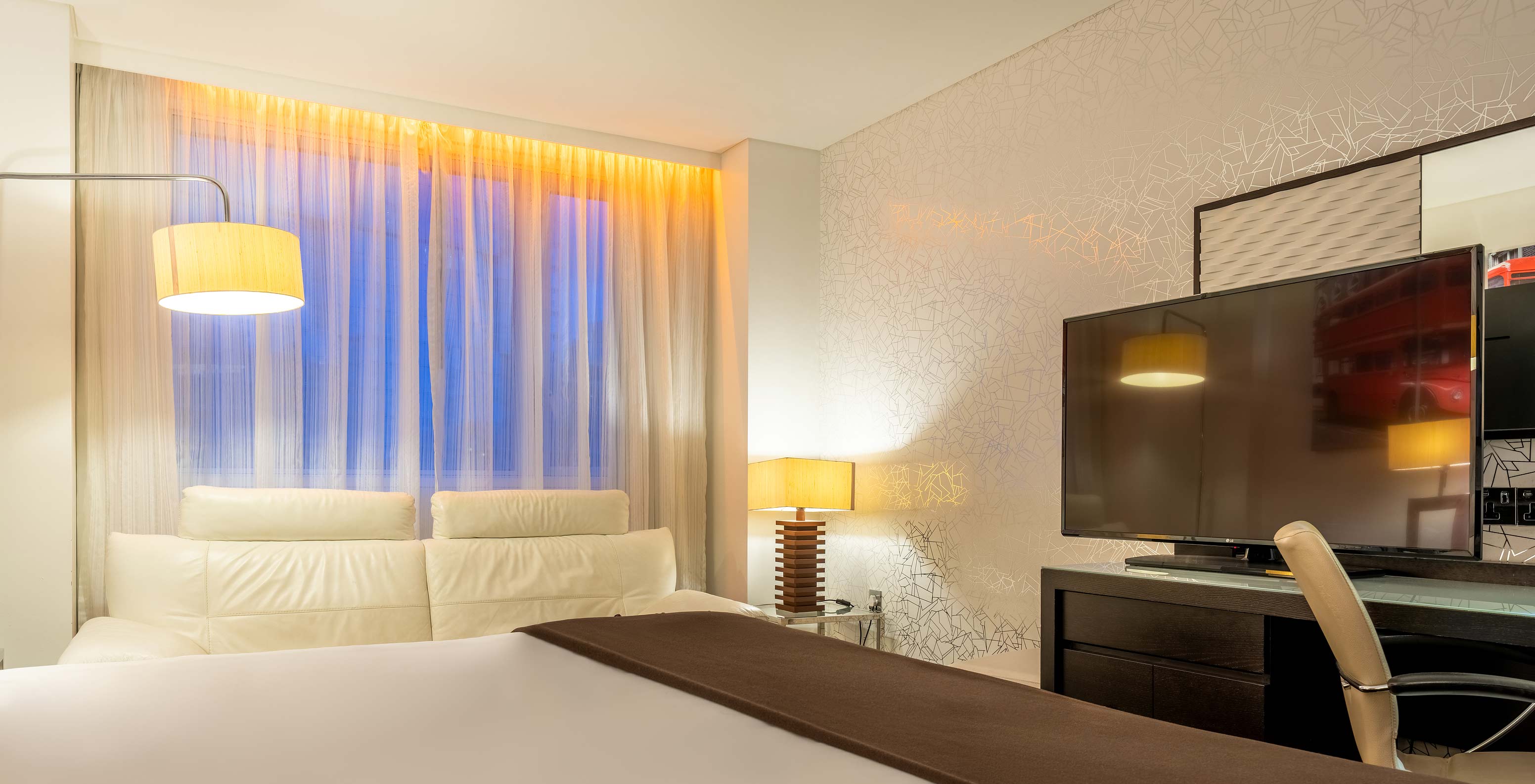 O Deluxe King do Pestana Chelsea Bridge tem cama, uma secretária com televisão e cadeira, sofá e janelas para a cidade