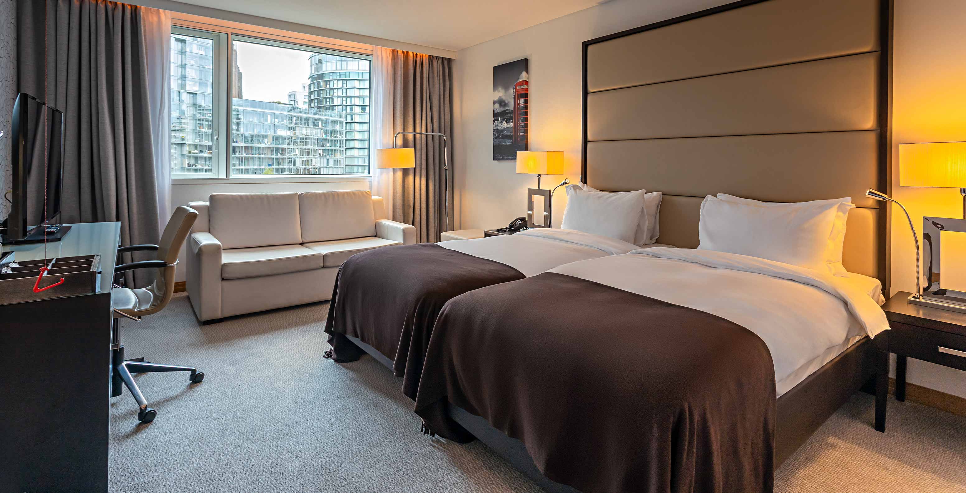 Quarto Standard Adaptado do Pestana Chelsea Bridge com duas camas, sofá e ampla janela urbana iluminada