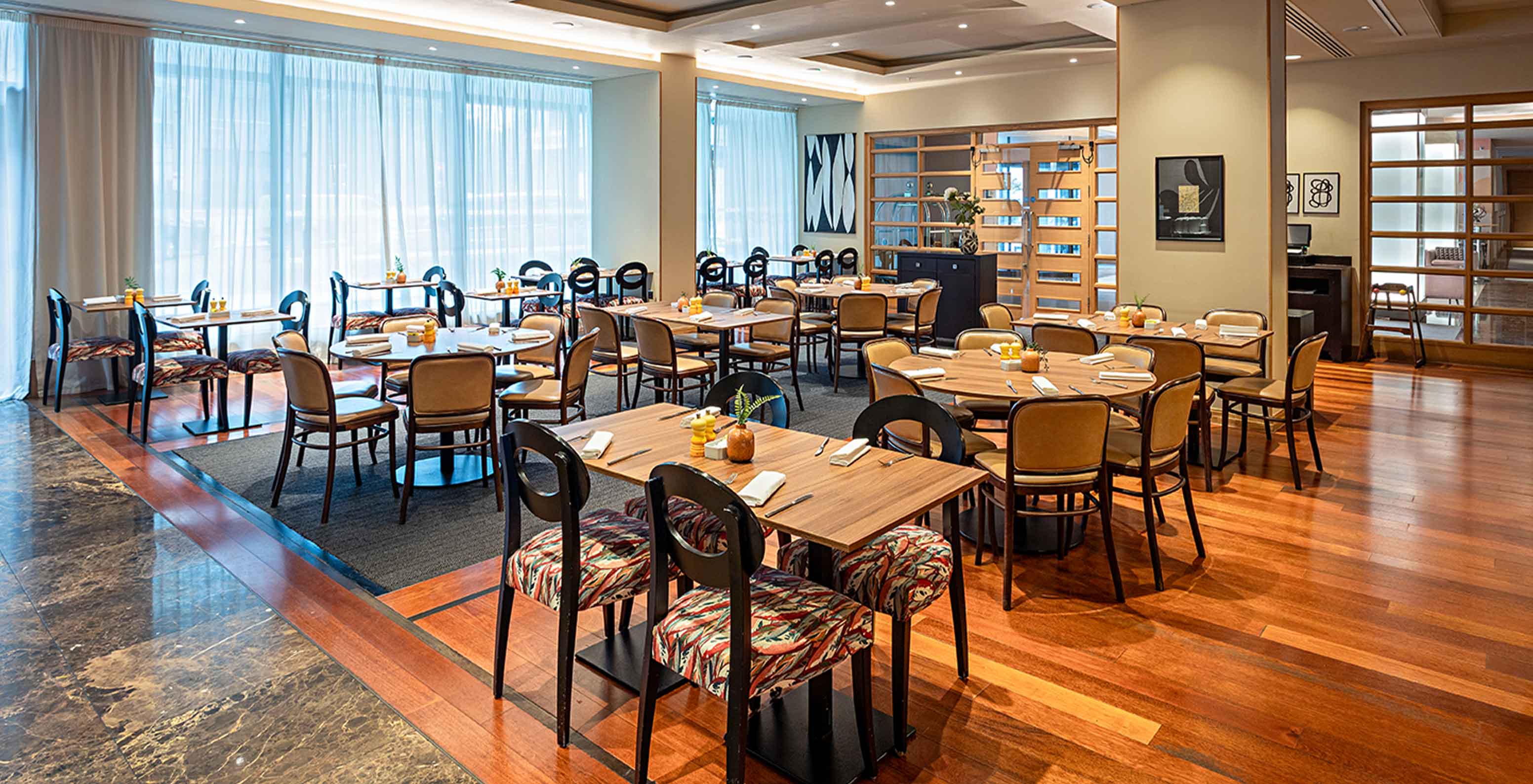 Sala de restaurante ampla com mesas postas, cadeiras em madeira, iluminação suave e janelas altas com cortinas