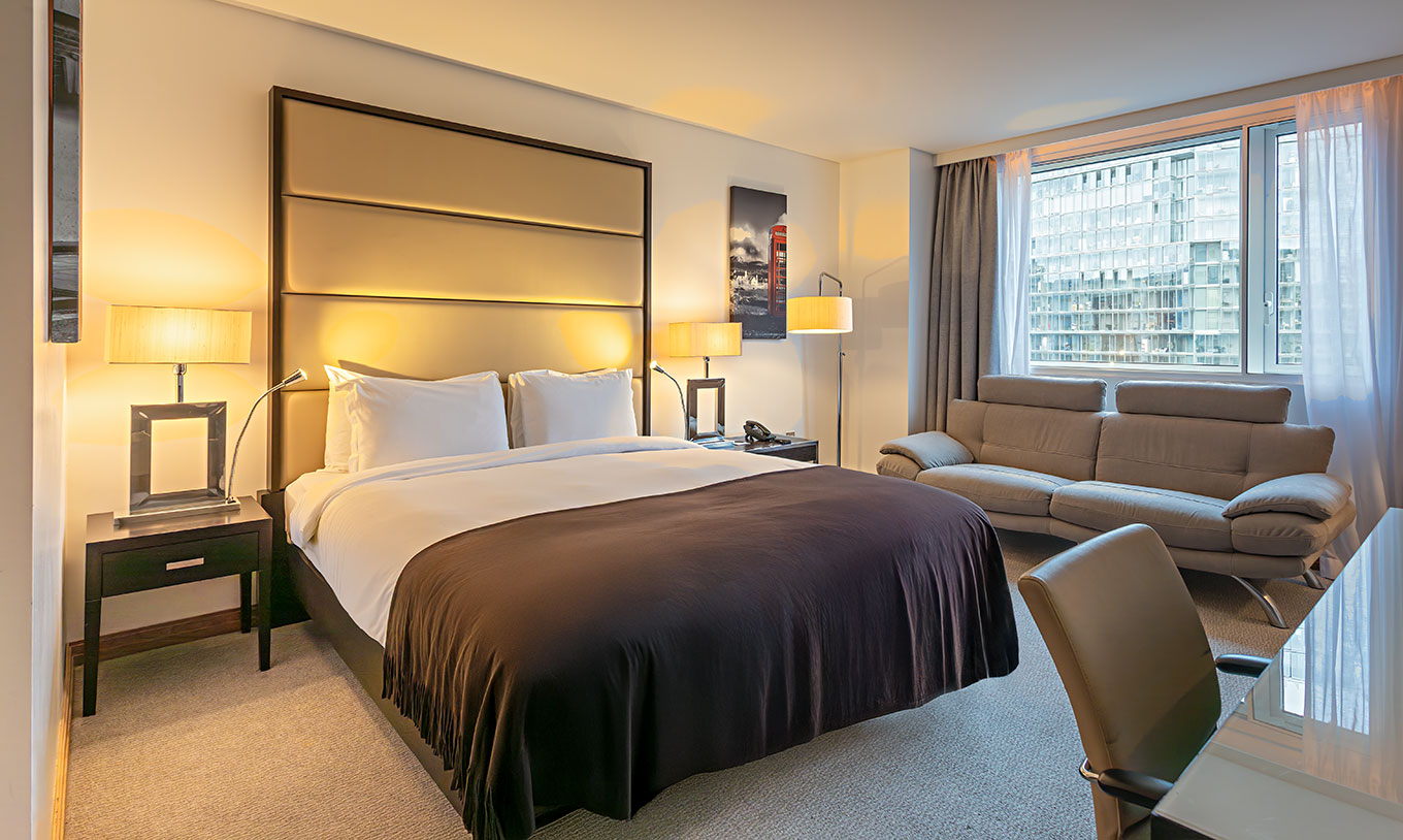 Quarto Deluxe King do Pestana Chelsea Bridge com cama de casal, sofá cinzento e janela ampla com vista urbana