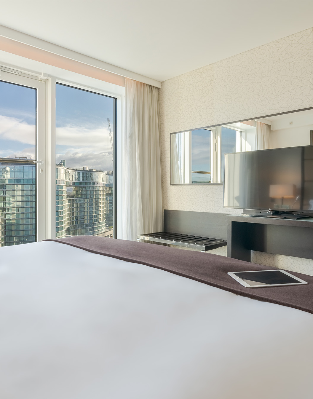 Quarto com cama de casal, televisão, espelho e janelas grandes com vista para a cidade de Londres