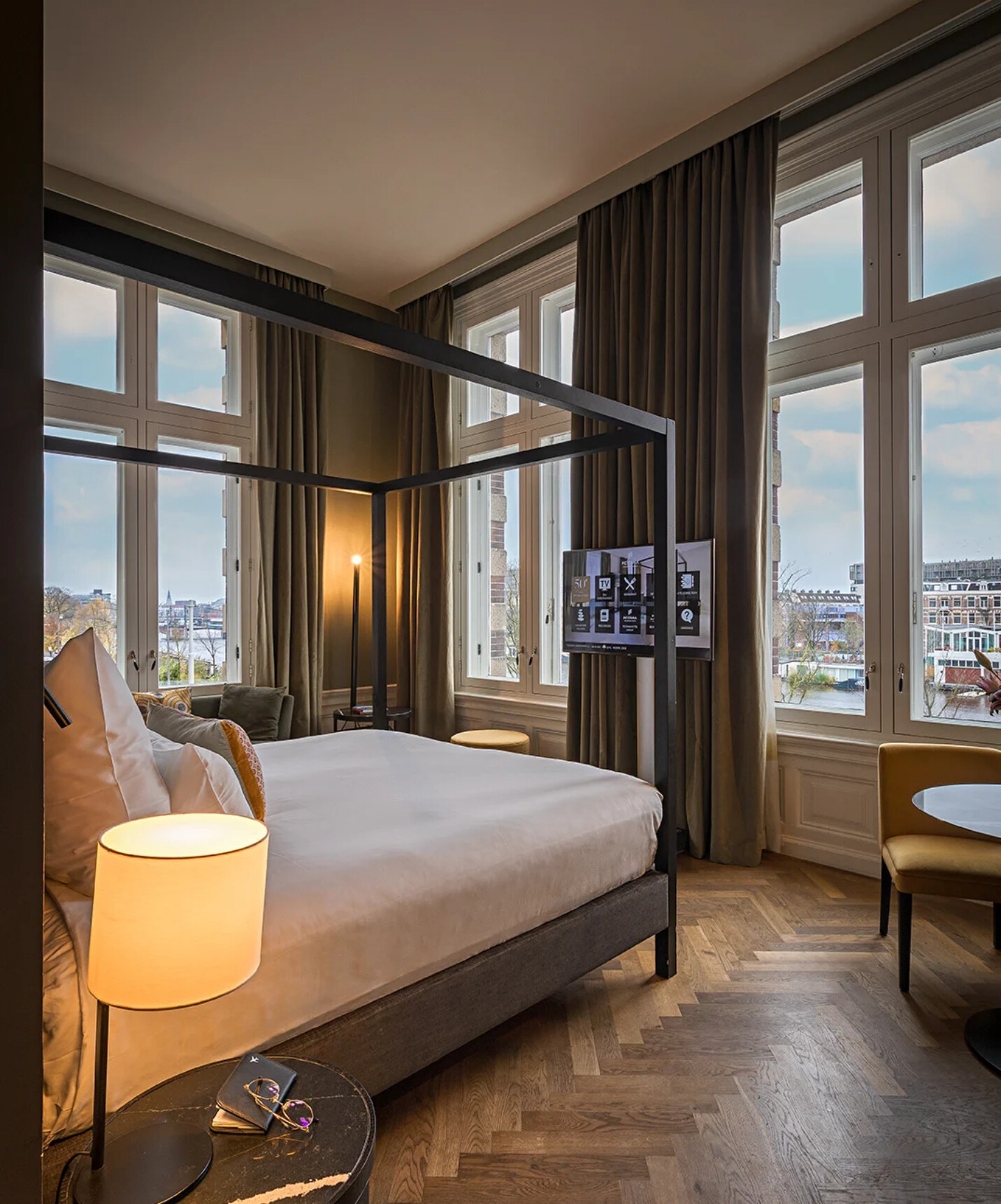 Quarto contemporâneo com cama de dossel, janelas amplas, cortinas escuras e vista urbana junto ao rio