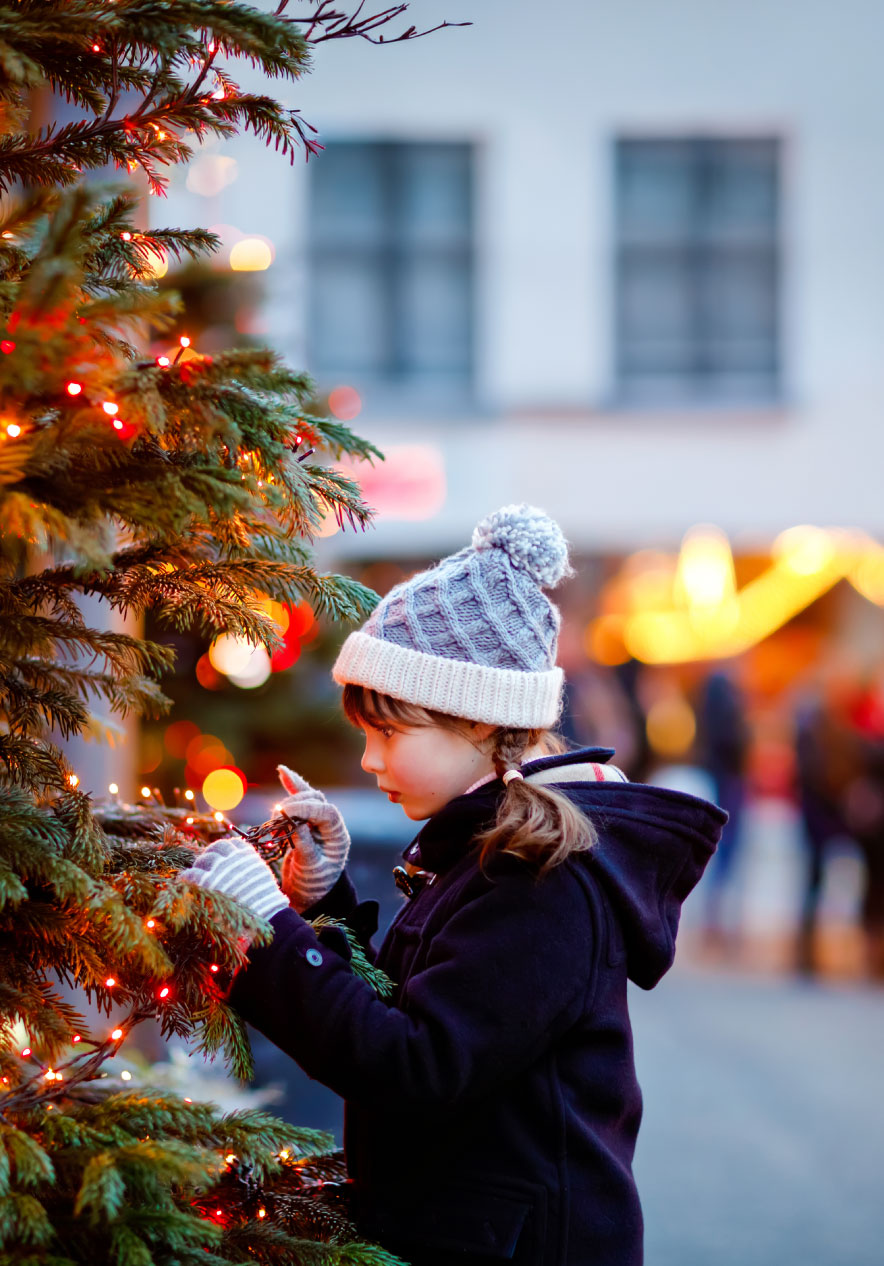 Criança de gorro e luvas junto a árvore de Natal iluminada num mercado natalício com luzes desfocadas