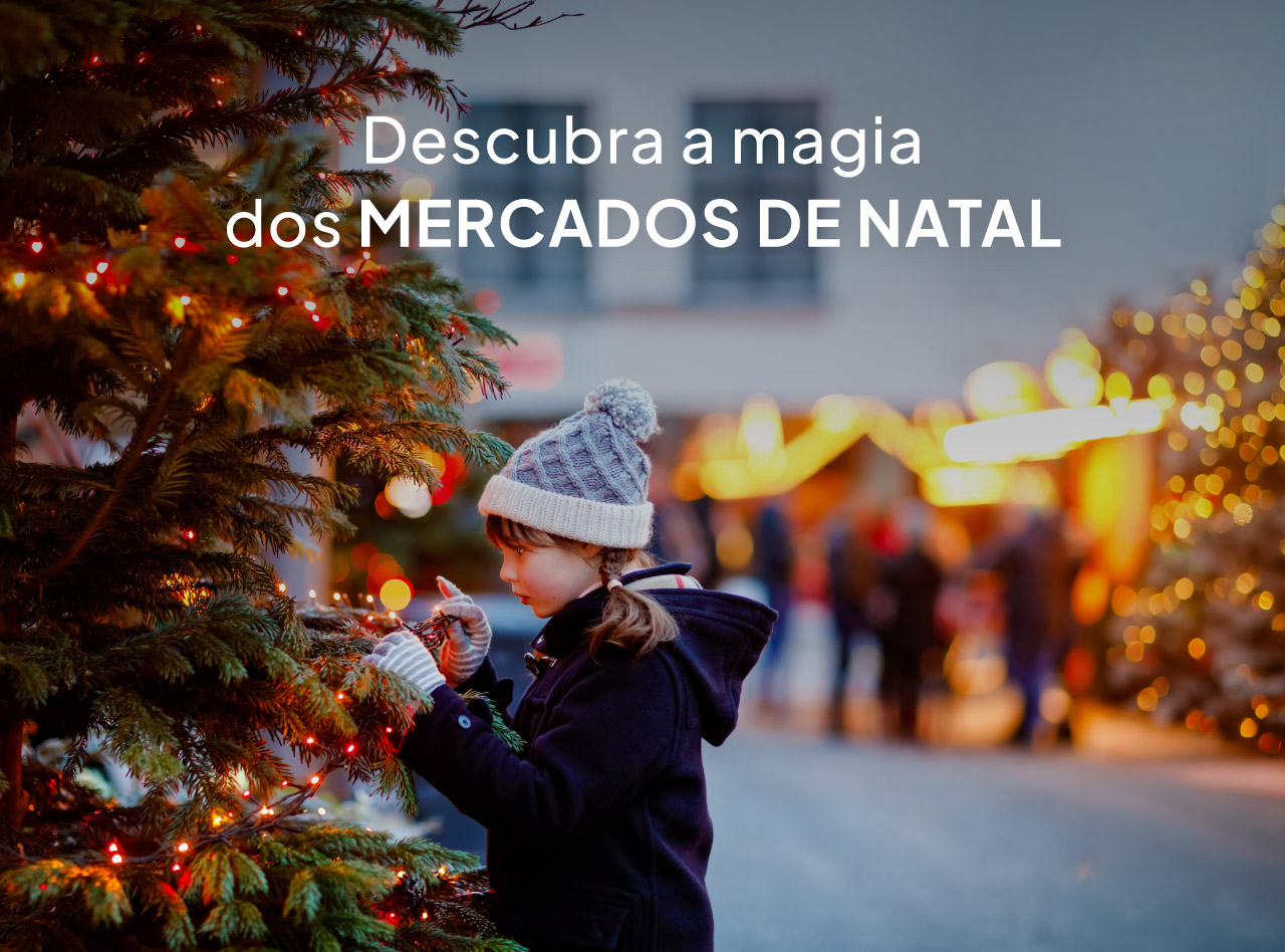 Criança junto a árvore iluminada num mercado de Natal com luzes decorativas e visitantes desfocados