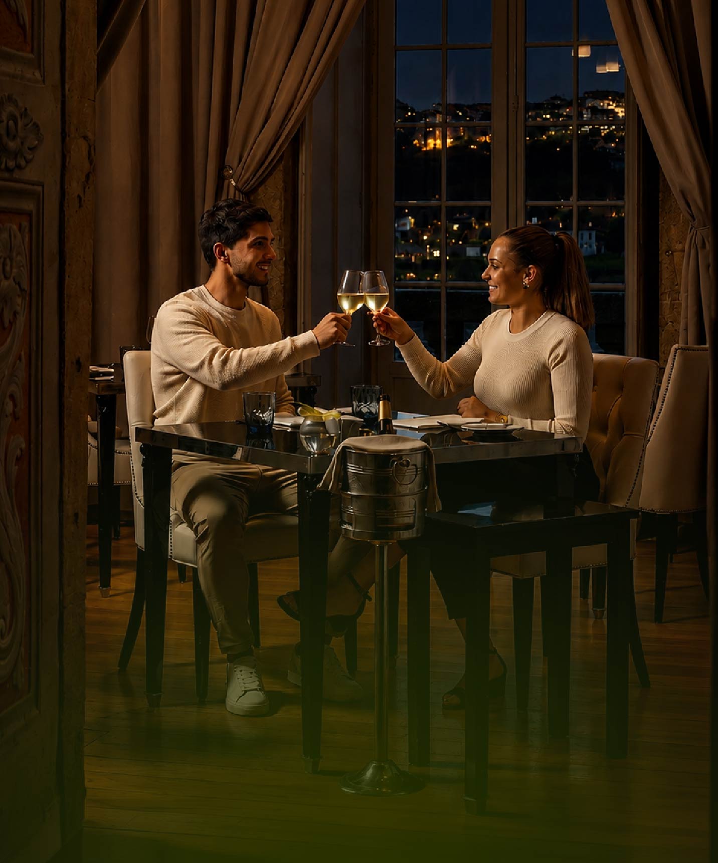 Casal brinda com vinho branco numa mesa junto à janela, com vista nocturna iluminada durante um jantar elegante