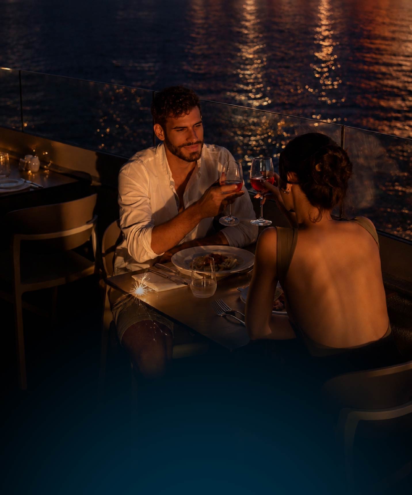 Casal brinda com vinho tinto durante um jantar à luz suave junto à água iluminada durante a noite tranquila