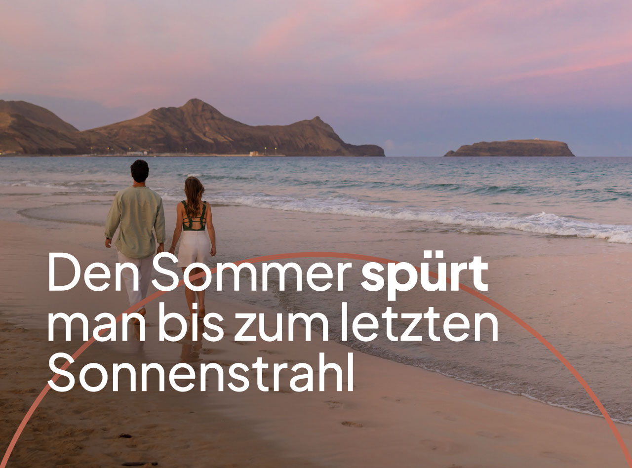 Paar spaziert am ruhigen Strand bei Sonnenuntergang mit Blick auf Berge, Meer und rosafarbenen Himmel