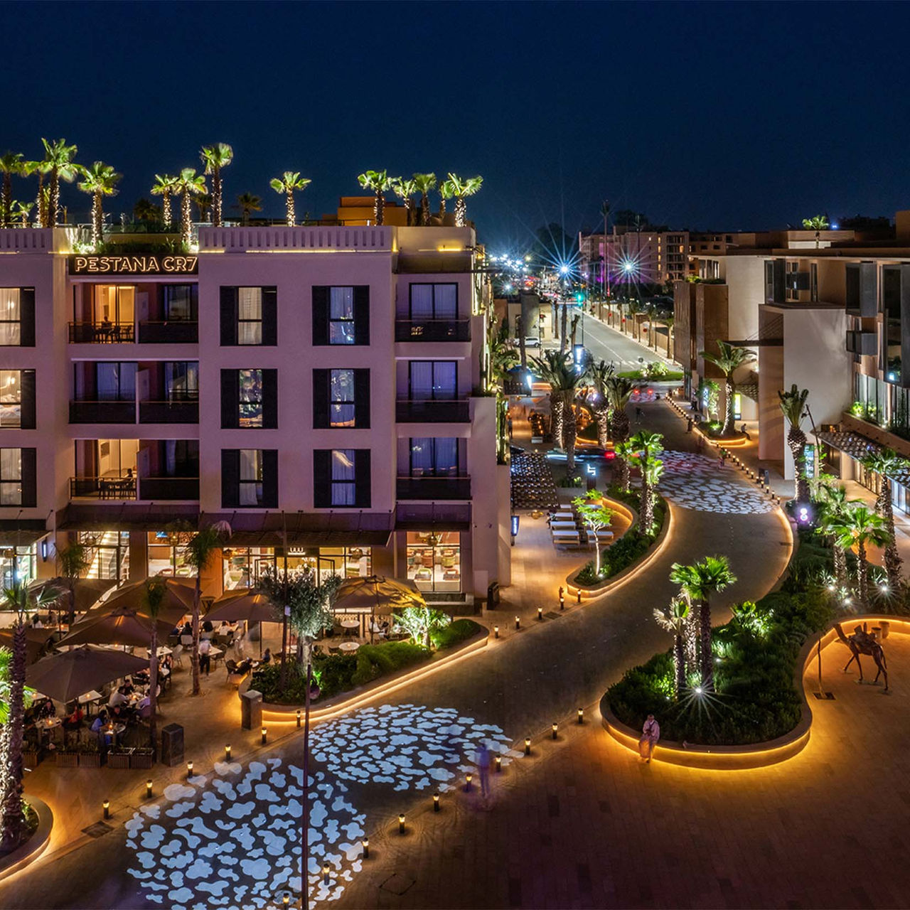 Außenansicht des Pestana CR7 Marrakech bei Nacht, in der M Avenue mit Geschäften, Palmen und beleuchteten Büschen