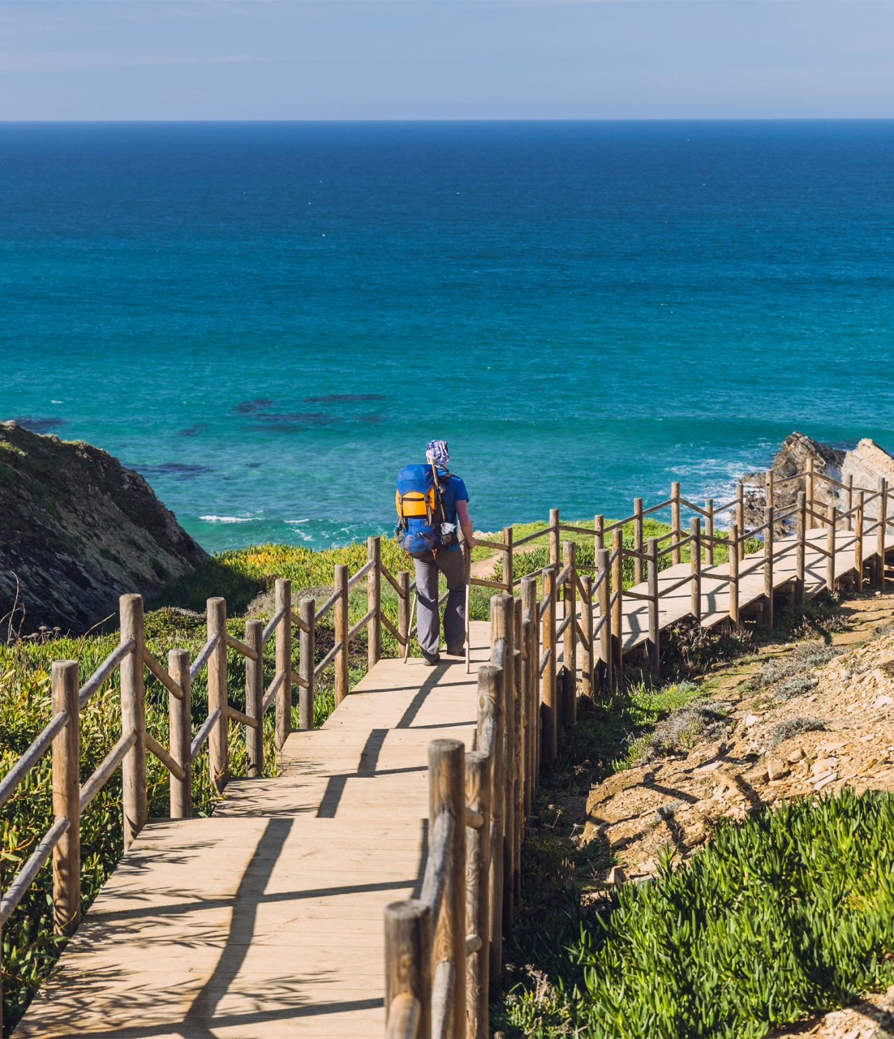 Mann geht entlang eines Holzstegs zu einem Strand in Alentejo, mit kristallklarem Wasser