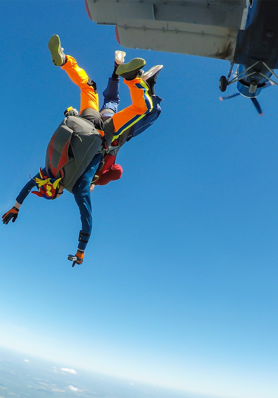 Zwei Personen springen mit Fallschirmen und bunten Anzügen aus einem Flugzeug in einen klaren Himmel
