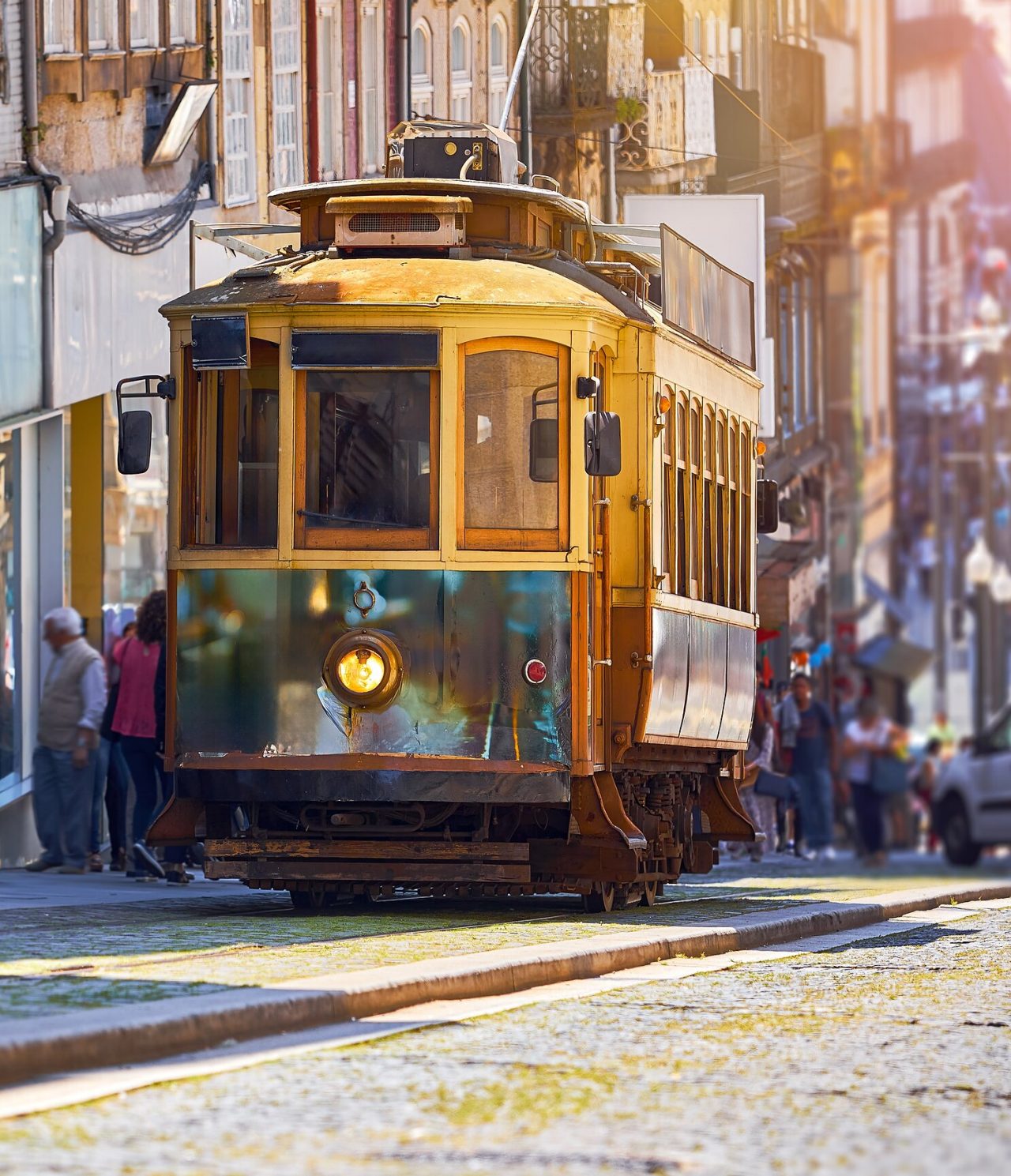 Blick auf eine alte gelbe Straßenbahn, die eine Straße in Porto hinauffährt mit mehreren Personen dahinter und alten Gebäuden