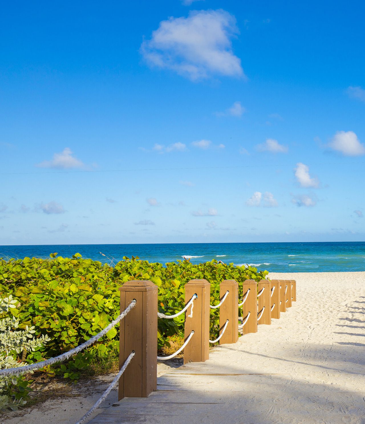 Weißer Sandstrand und kristallklares blaues Meer, mit grüner Vegetation und einem Holzsteg zum Strand