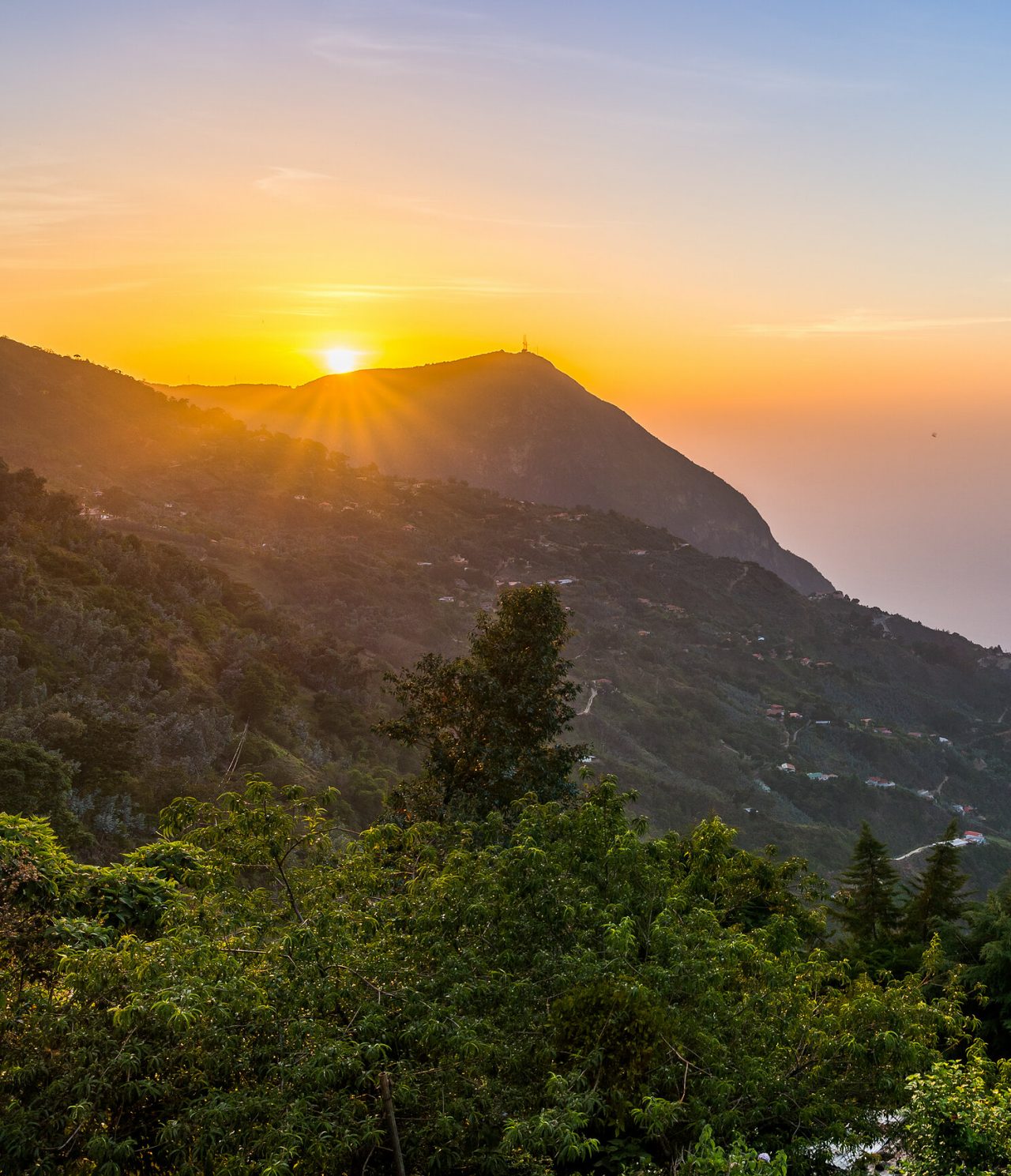 Luftaufnahme eines Berges in Caracas, mit Vegetation und einigen Häusern, mit buntem Himmel im Hintergrund
