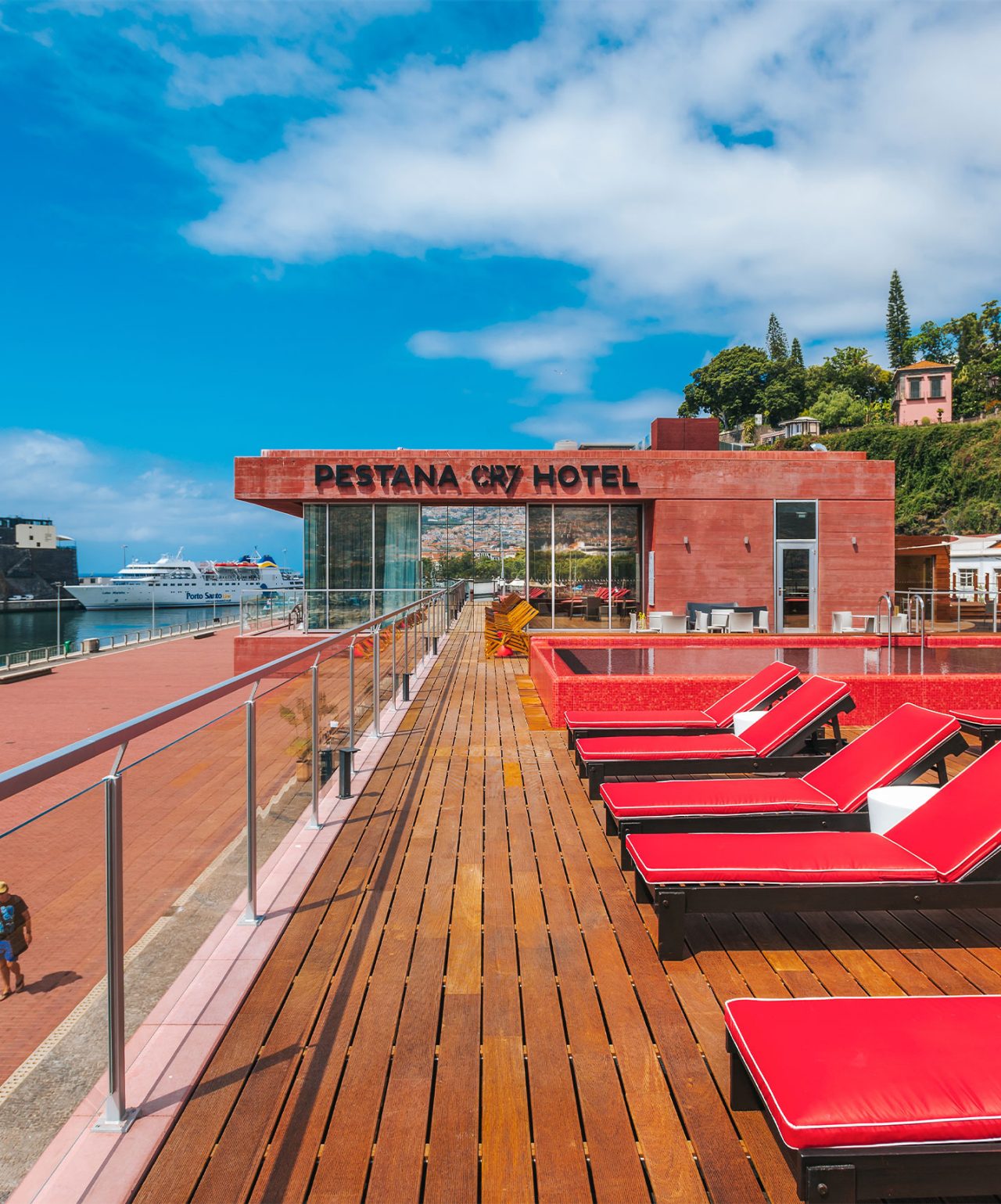 Die Marke Pestana CR7 Lifestyle Hotels mit roter Sonnenterrasse, Liegen und Blick auf den Hafen bei blauem Himmel