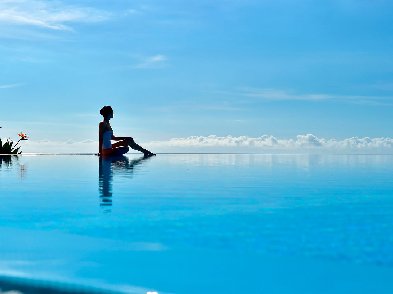 Mulher sentada à beira de uma piscina infinita ao ar livre com vista para o horizonte azul e nuvens
