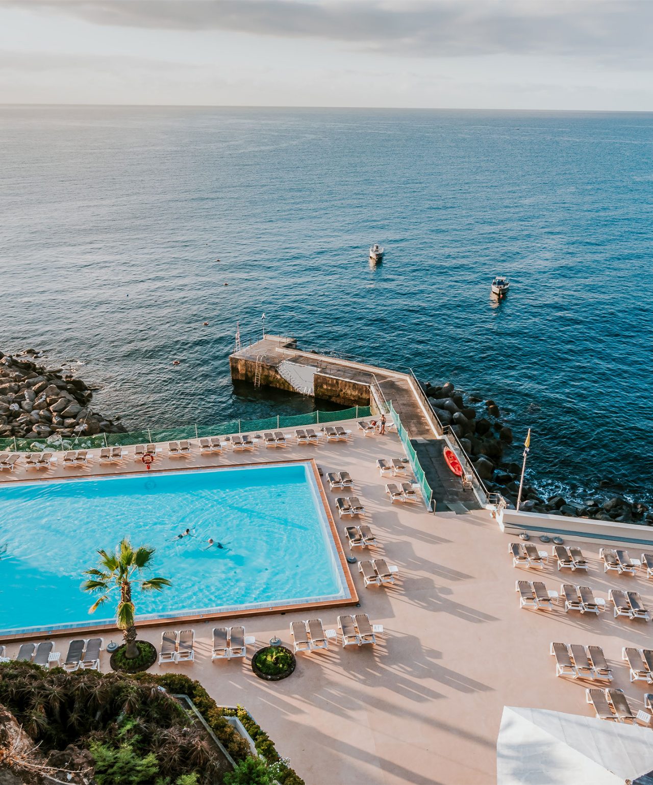 Piscina exterior com espreguiçadeiras junto ao mar, vista para o oceano e pequeno cais com barcos