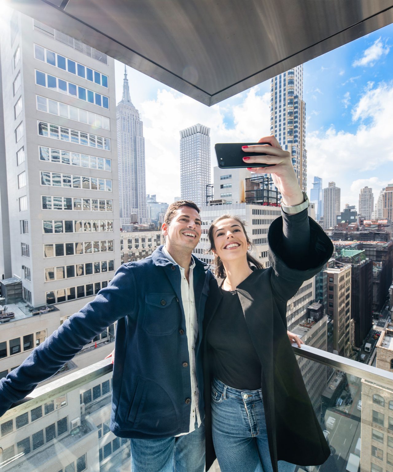 Casal tirando selfie em varanda com vista para prédios altos e céu azul em dia ensolarado