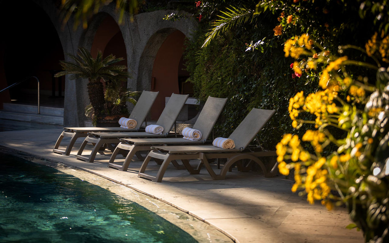 Sonnenliegen mit gerollten Handtüchern am ruhigen Pool neben bepflanzten Bögen und gelben Blüten im Schatten