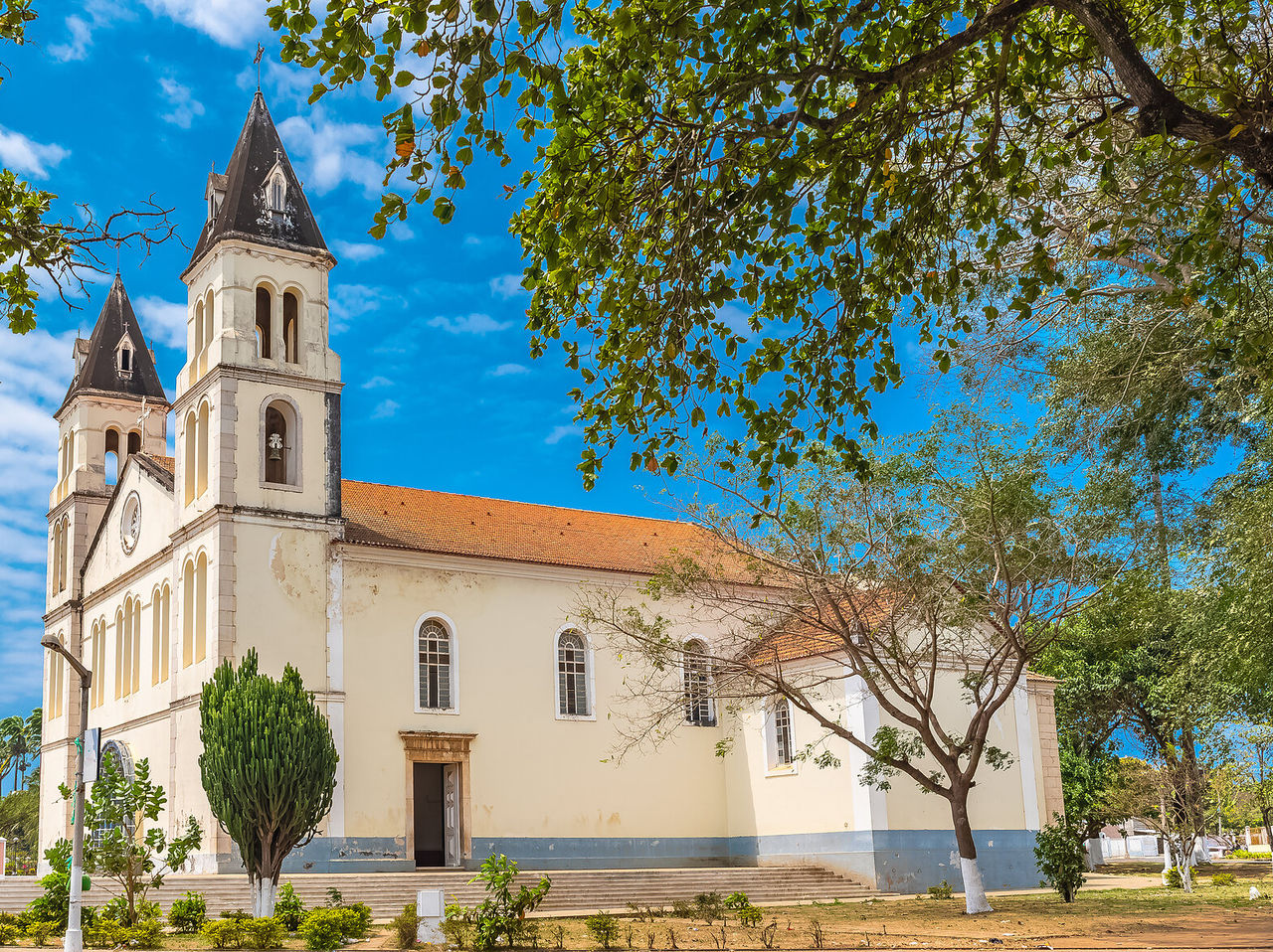 Explore Hotels in São Tomé featuring a historic white church with twin towers beneath leafy trees and blue sky