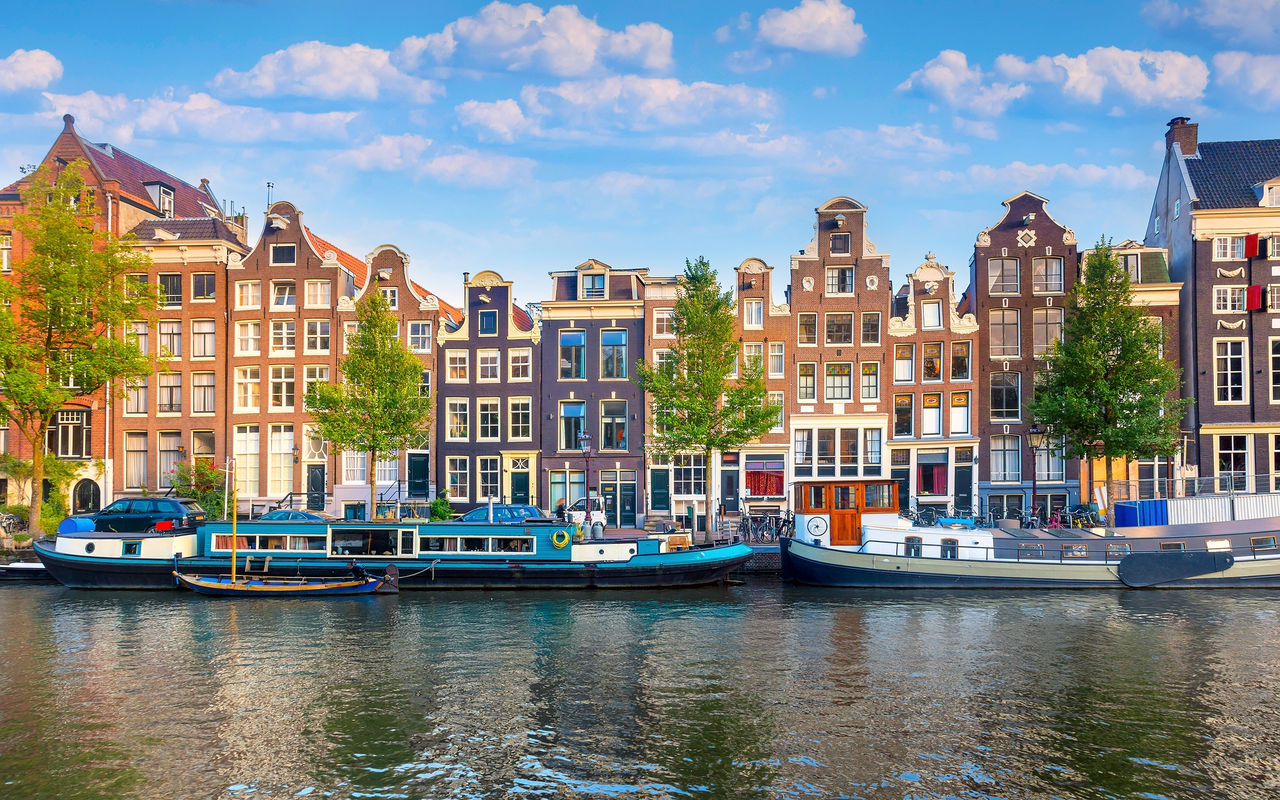 Amsterdam canal with people walking along the shore, boats, and typical buildings under a blue sky