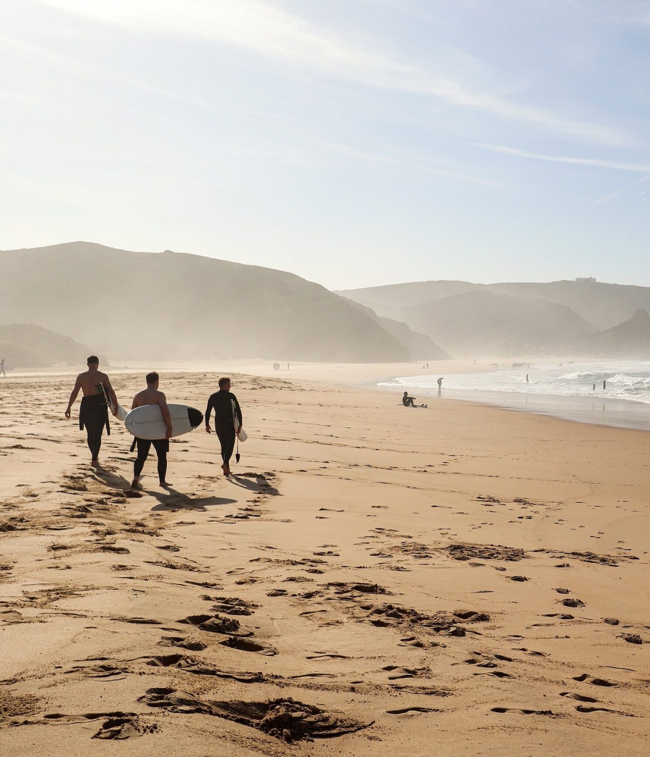 Explore Hotels in the Algarve featuring surfers walking along a misty beach beneath coastal cliffs