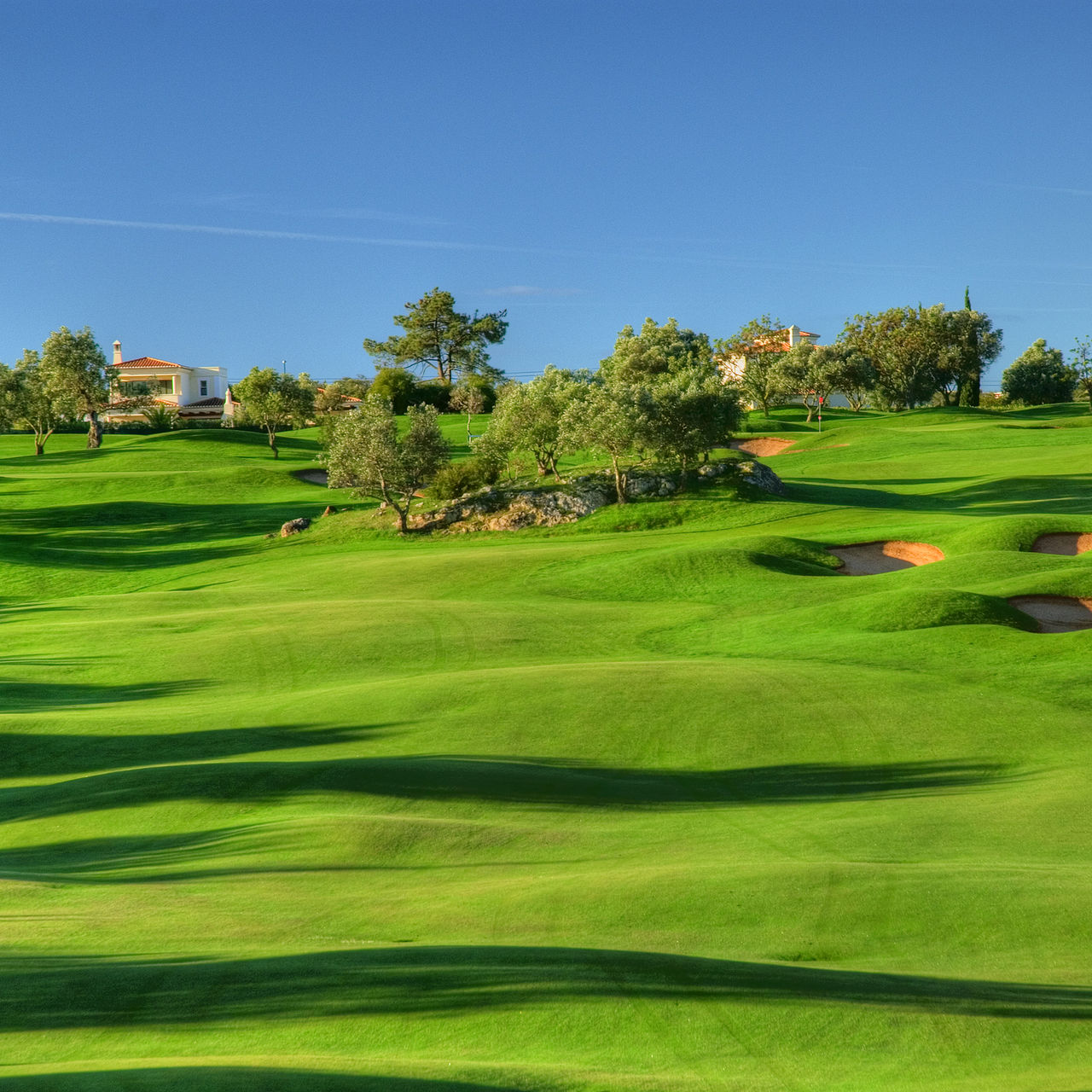 Lush golf course in rolling terrain at Gramacho, with blue sky, trees, and bushes, and some houses in the background