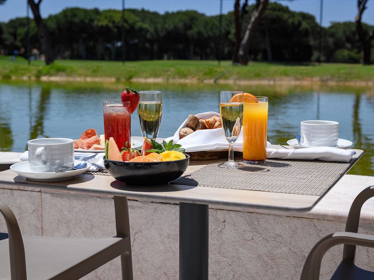 Restaurant at Pestana Vila Sol, Hotel with Golf and Spa in the Algarve, featuring a table set with fresh fruit, juices, and pastries beside a tranquil lake under clear blue skies