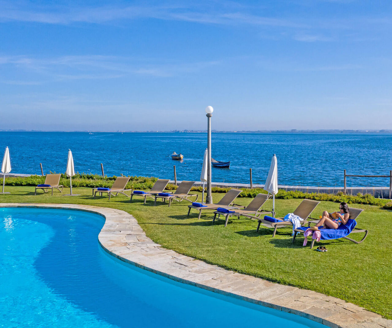 center-portugal hotel overlooking a curved pool, sun loungers on grass and calm blue sea beyond under clear skies