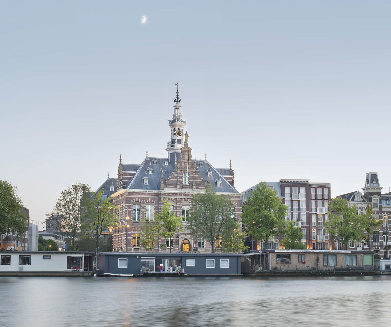Amsterdam hotel overlooking a historic canal building at dusk with houseboats, trees and a crescent moon