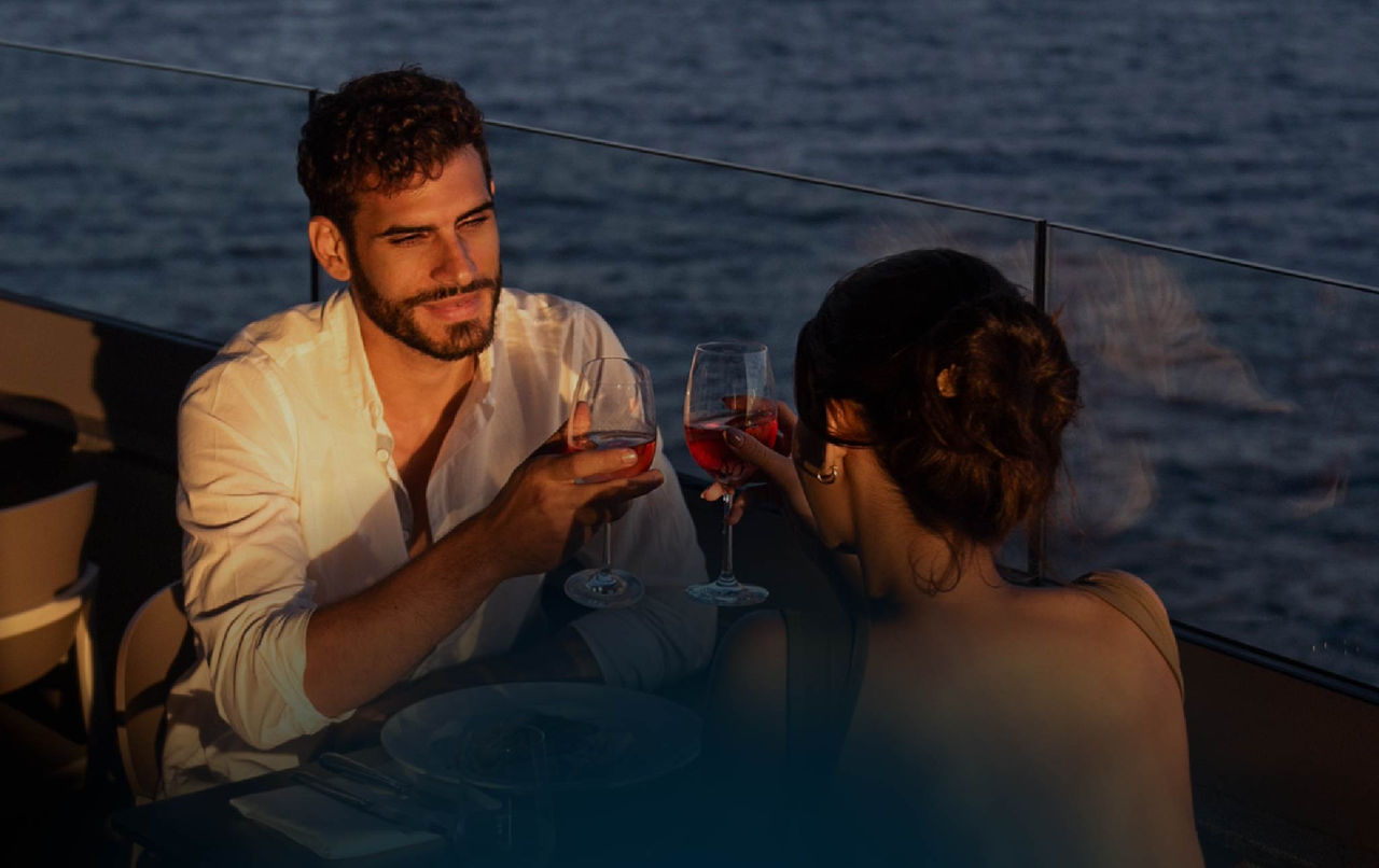 Couple raising glasses during a sunset dinner beside the water on an outdoor terrace with warm evening light