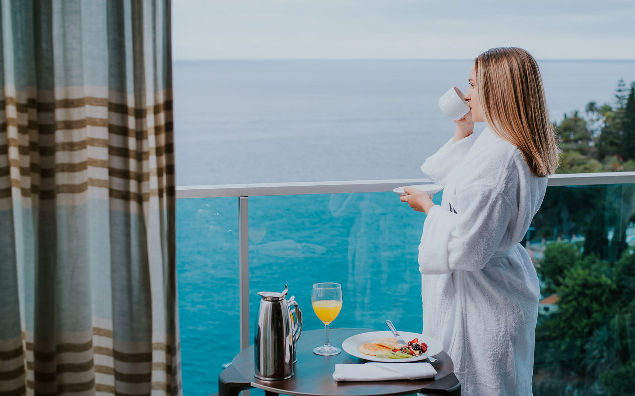 Mulher de roupão bebe café numa varanda com vista para o mar, mesa com pequeno-almoço e sumo de laranja