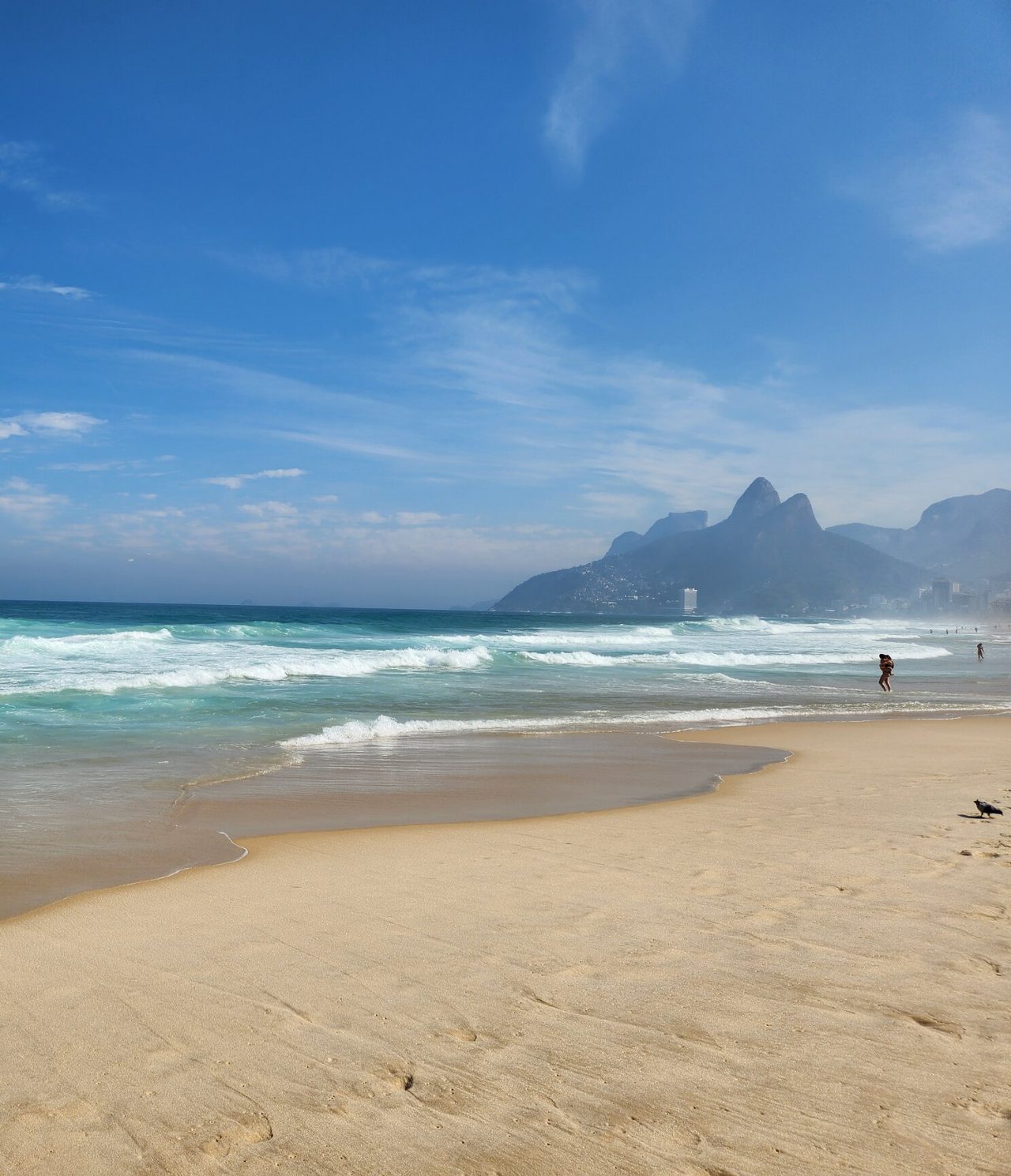 Vista de una playa en Río de Janeiro, con arena blanca, agua turquesa, montañas y personas al borde del agua