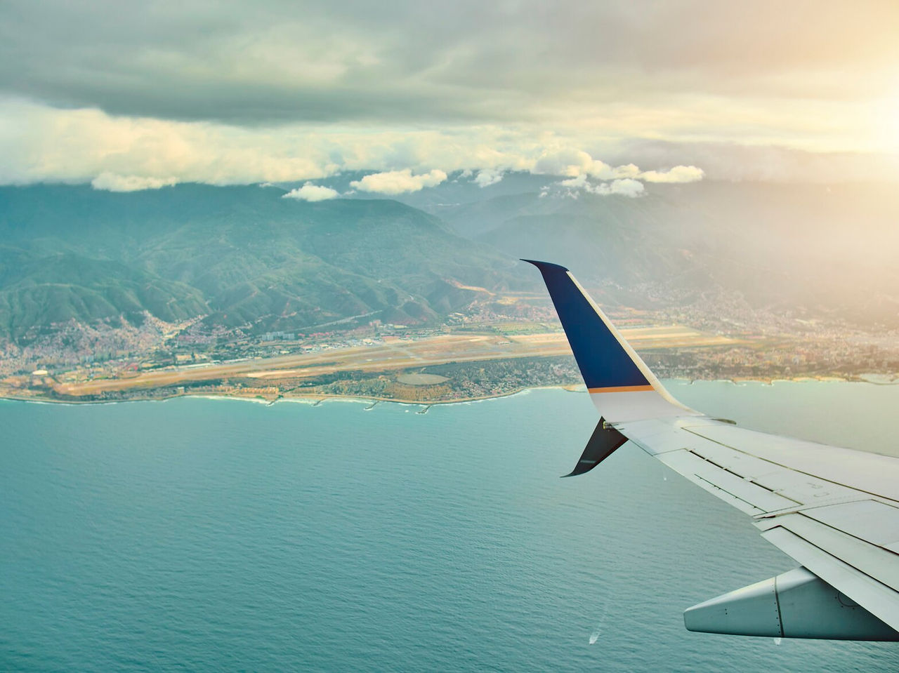 Vista aérea del ala de un avión sobrevolando un océano, con una ciudad al fondo, rodeada de montañas