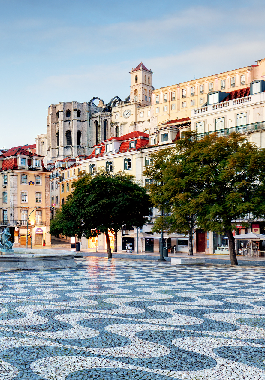 Vista de la Plaza del Rossio con calzada portuguesa, una fuente central con estatuas y varios edificios alrededor