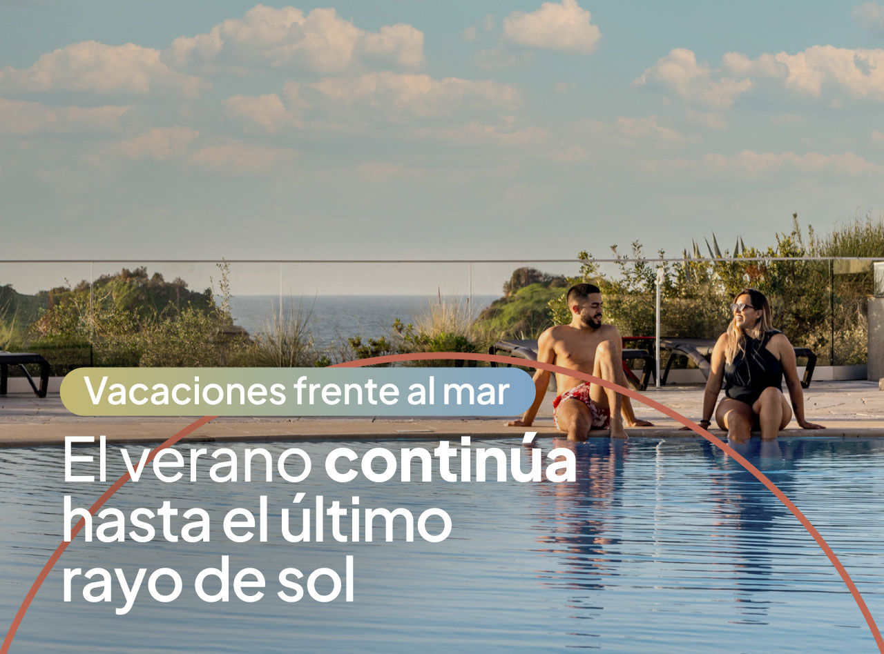 Pareja sentada junto a una piscina infinita con vistas al mar bajo un cielo azul con nubes al atardecer