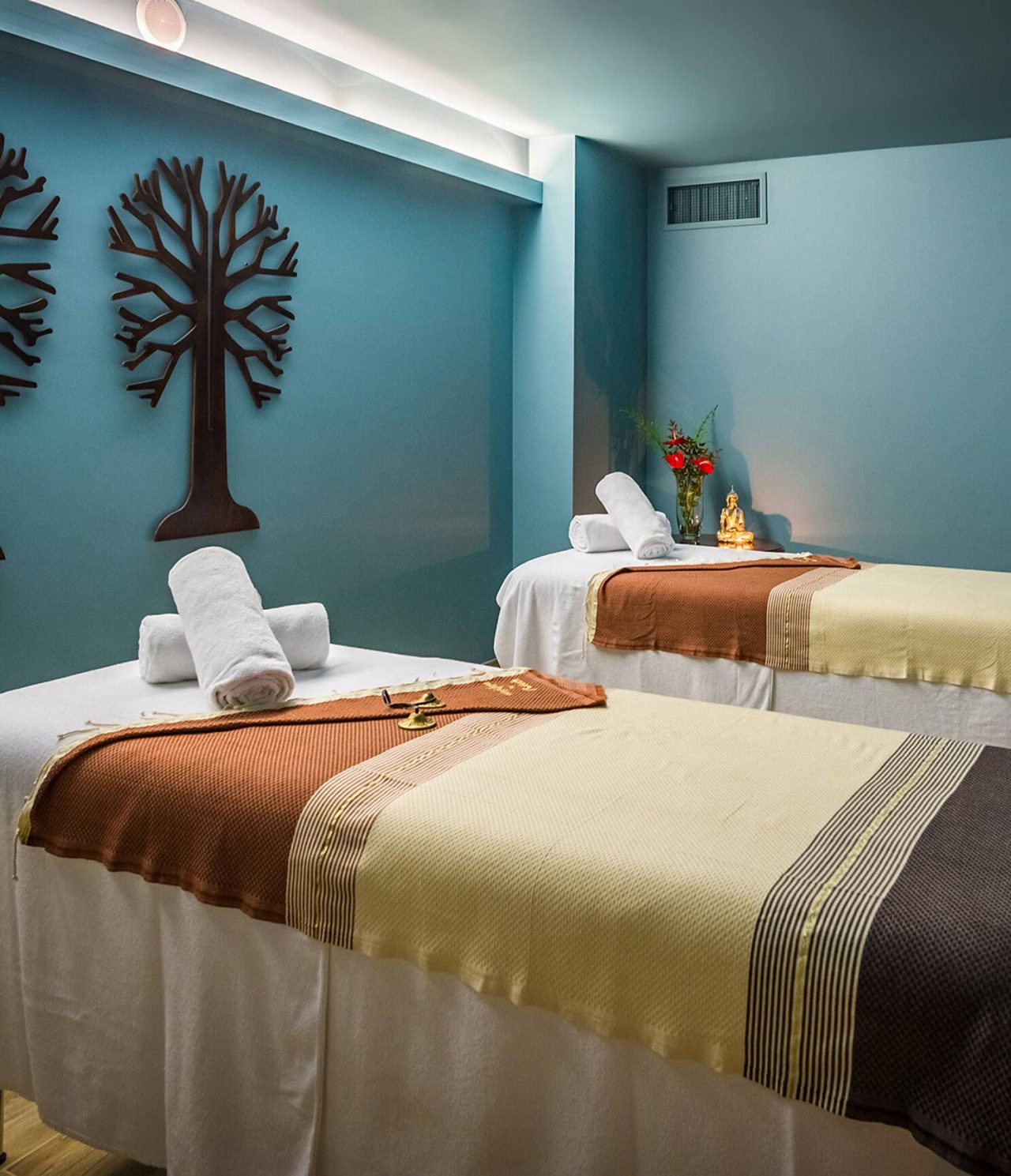 Sala de spa con dos camillas de masaje, toallas enrolladas y decoración tenue sobre paredes azul verdoso