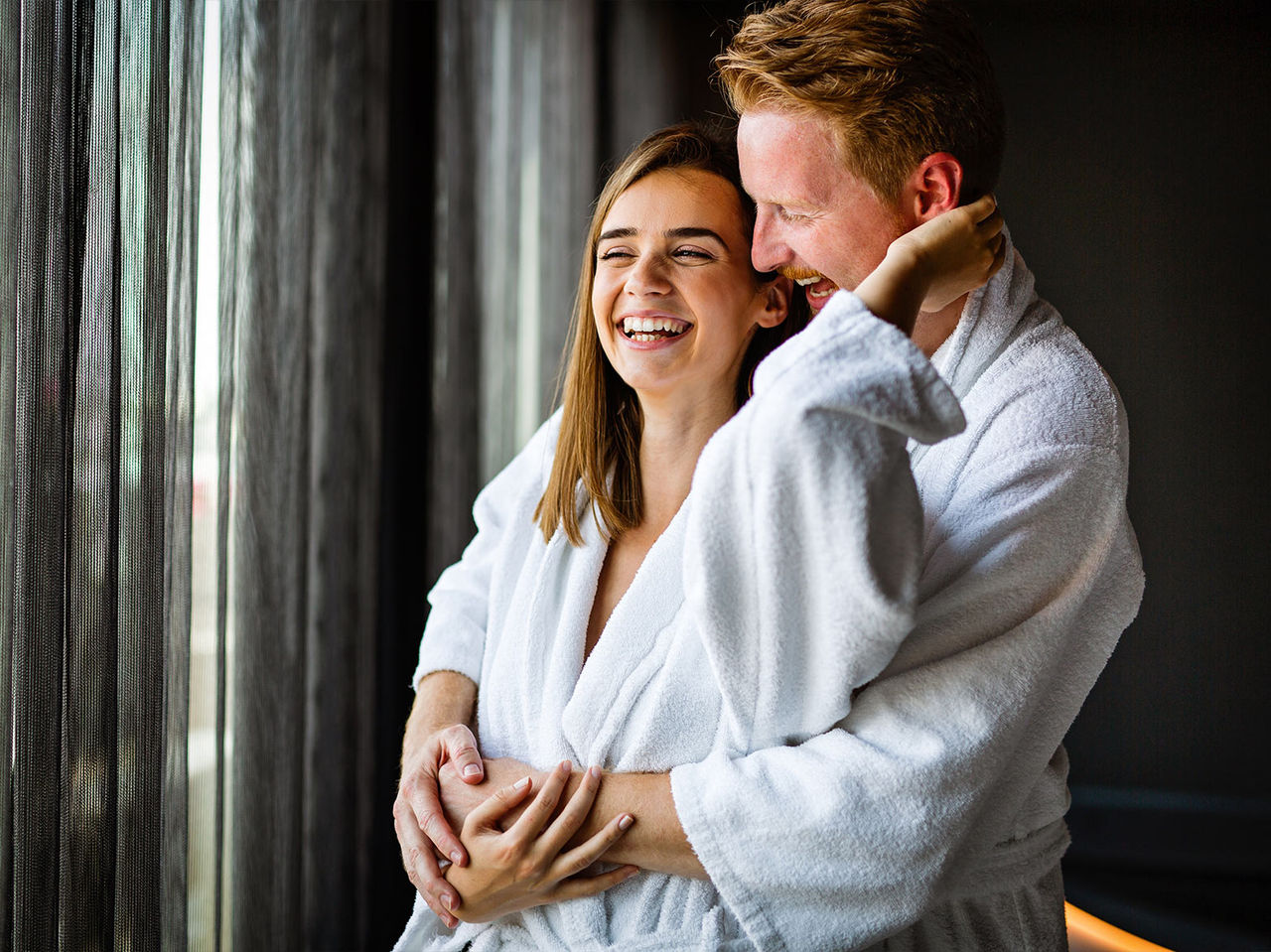 Pareja con albornoces blancos sonriendo junto a una ventana en un ambiente relajante de spa interior