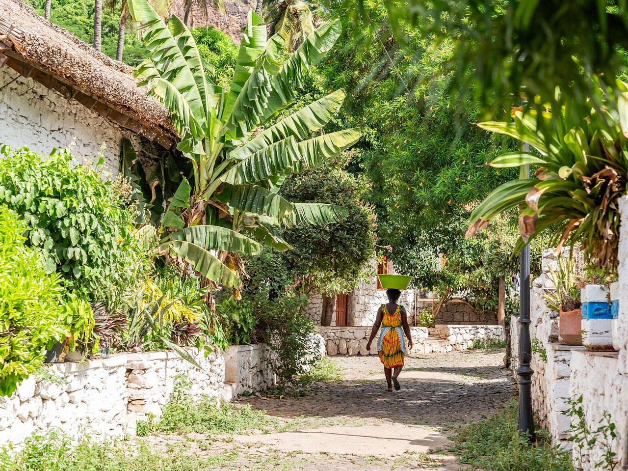 Femme cap-verdienne marchant dans une rue étroite avec une bassine sur la tête, entourée de végétation tropicale