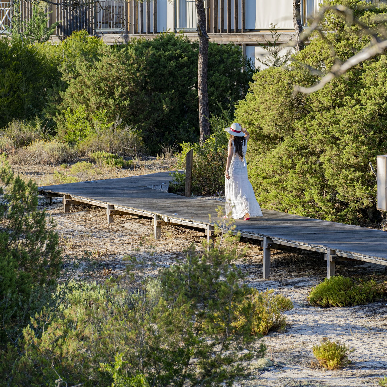 Passerelle en bois au Pestana Tróia Eco Resort, où une femme en robe et chapeau se promène entre les villas