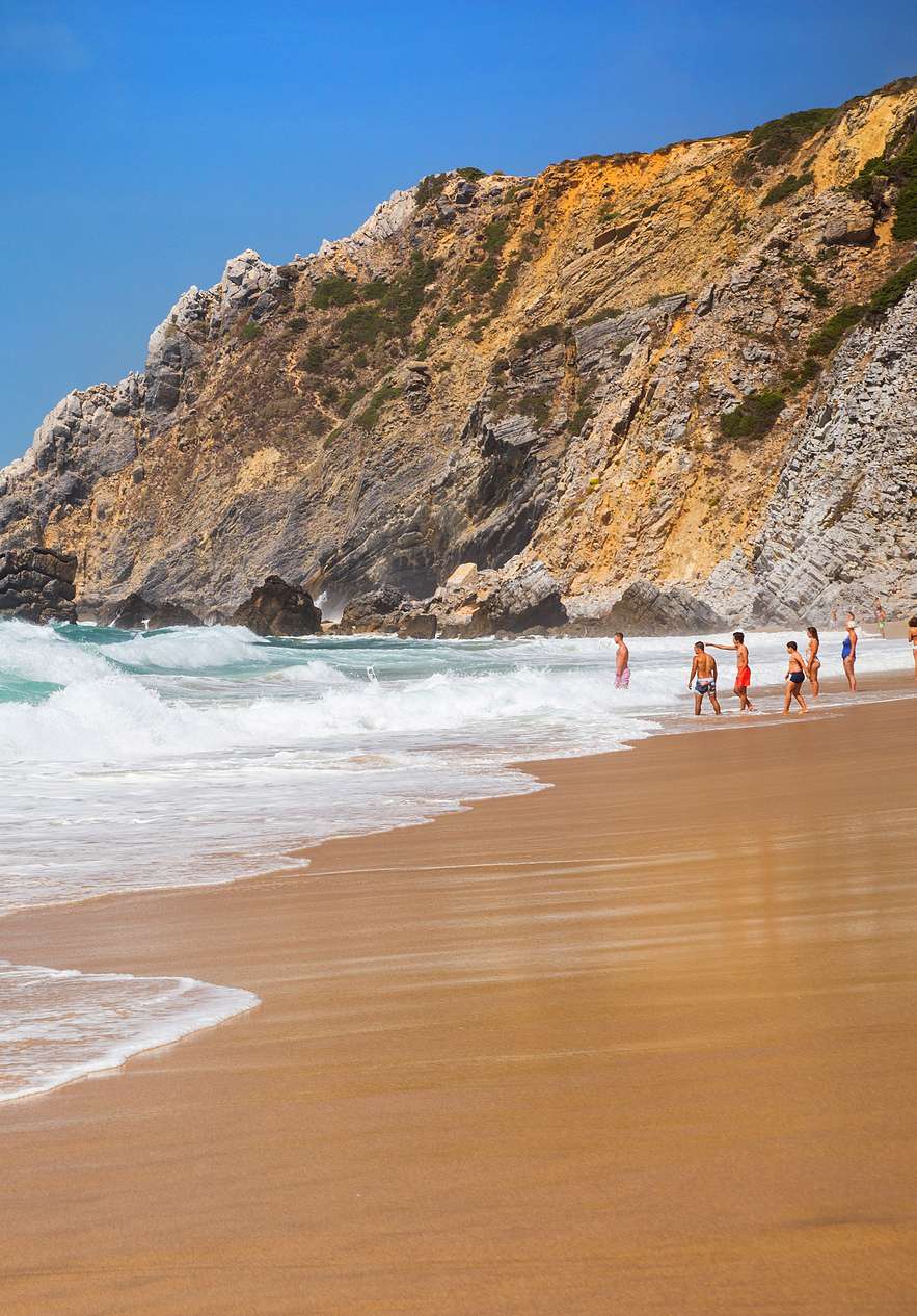 Plage à Sintra, avec du sable doré et des vagues se brisant sur la côte, des gens entrant dans l'eau et des rochers