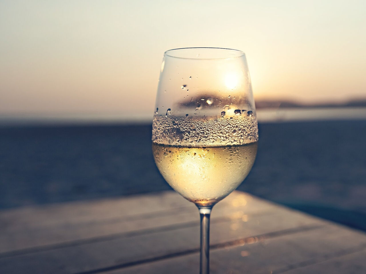 Verre de vin blanc avec des reflets flous du coucher de soleil en arrière-plan, sur une surface en bois