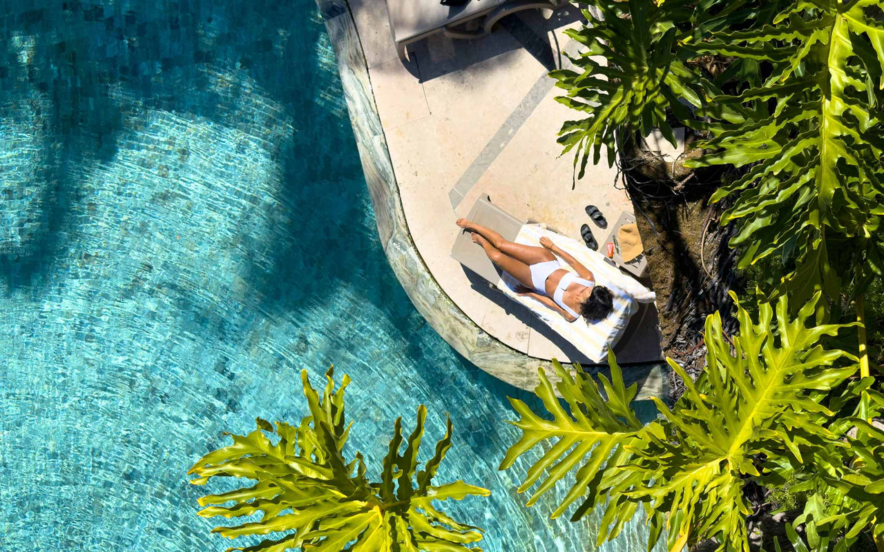 Femme allongée sur une chaise longue au bord d'une piscine, entourée de palmiers et de végétation tropicale