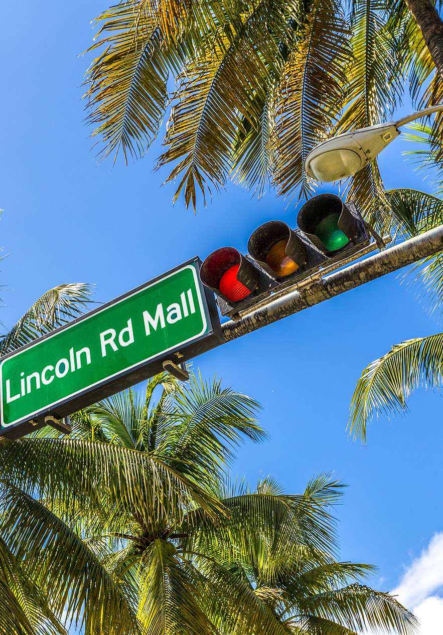 Verkeersbord voor "Lincoln Rd Mall" met stoplicht ernaast, omringd door palmbomen en een helderblauwe lucht